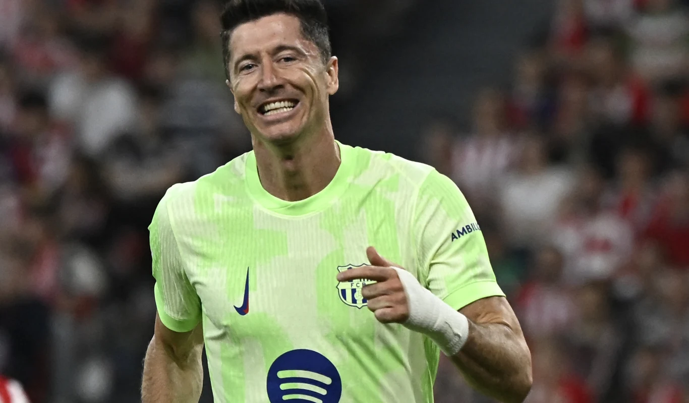 Robert Lewandowski Piłkarz w neonowym stroju drużyny FC Barcelona z uśmiechem na twarzy wykonuje gest palcem, z widocznym logo sponsora na koszulce, w tle rozmyta publiczność stadionowa.