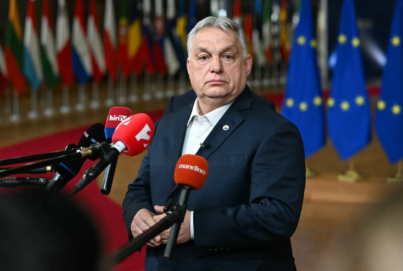 Viktor Orbán Viktor Orbán w garniturze przed mikrofonami mediów, w tle flagi państwowe i Unii Europejskiej
