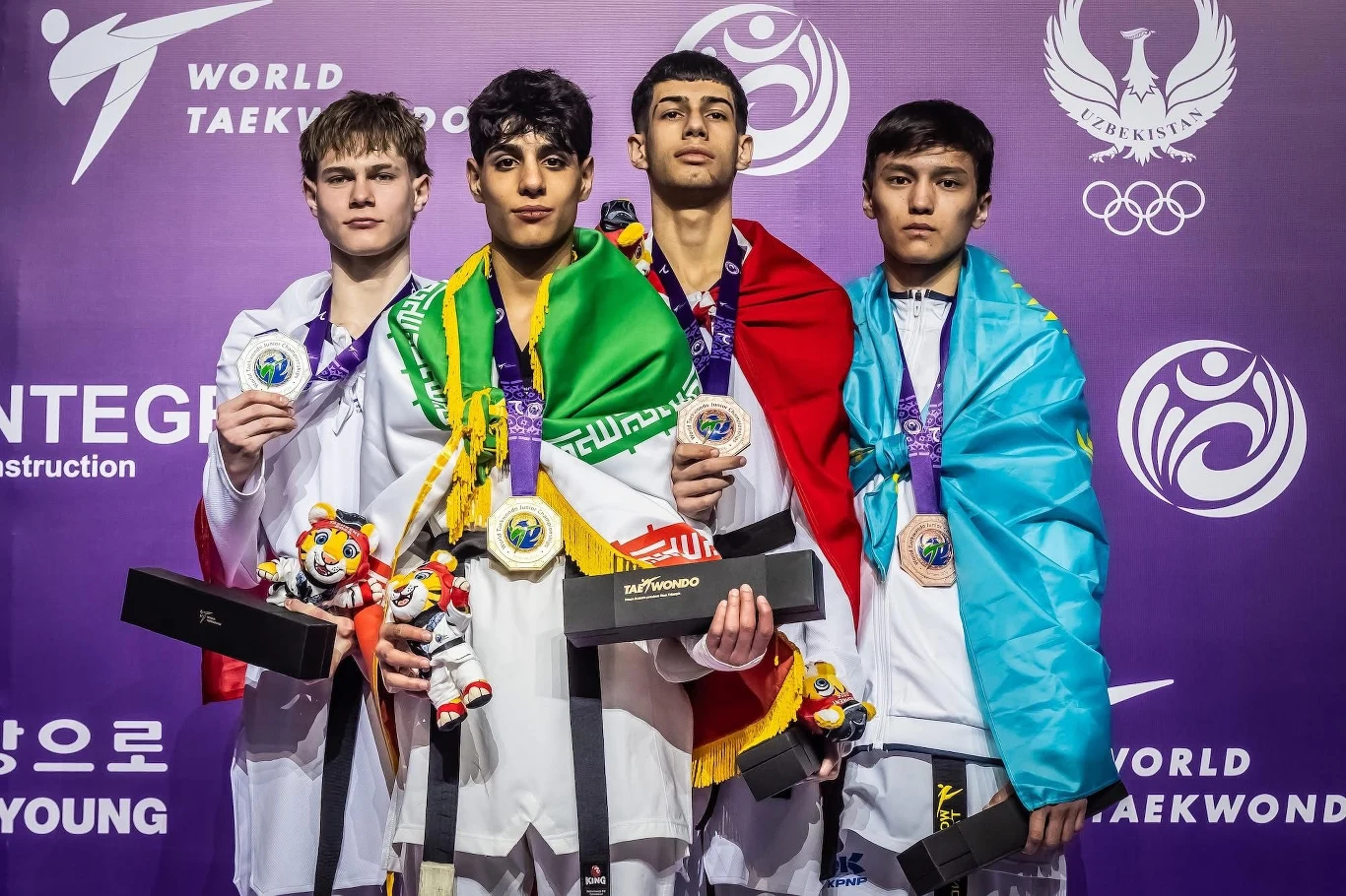 Czterech młodych sportowców w strojach taekwondo stoi na podium z medalami na szyjach, każdy z nich trzyma maskotkę turniejową oraz upominki, a wyróżniają się flagami krajów zawieszonymi na ramionach, na tle fioletowej ścianki z logotypami mistrzostw i...