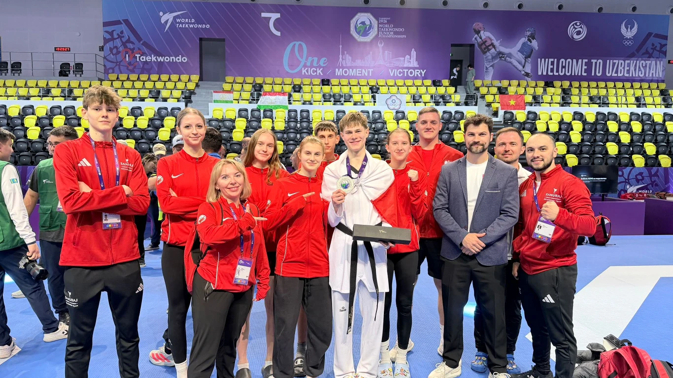 grupa osób ubranych w czerwone dresy i trenerzy pozuje na hali sportowej przy zawodniku w białym stroju taekwondo, który trzyma medal; w tle trybuny, banery oraz napis 'Welcome to Uzbekistan'