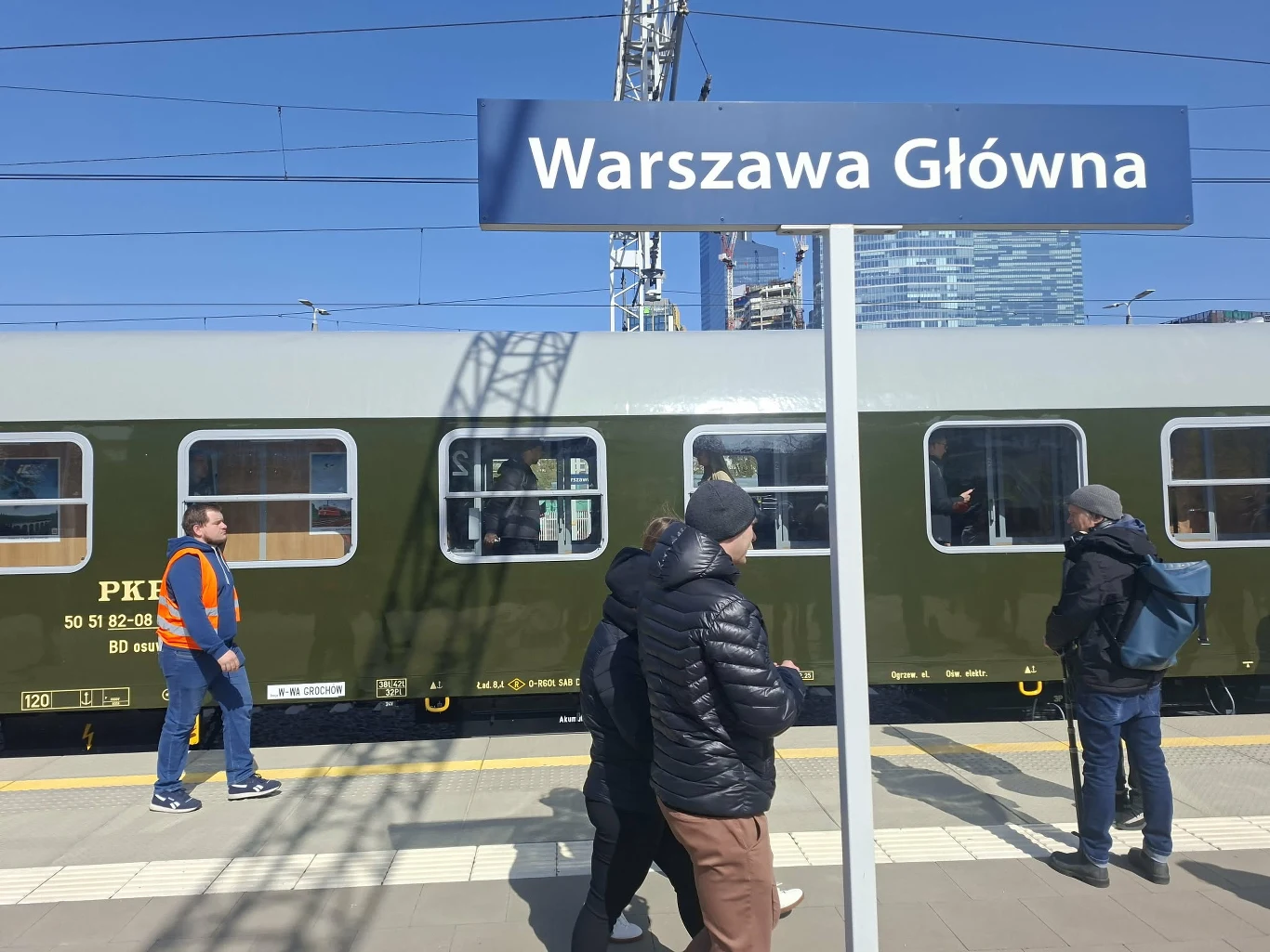 peron dworca kolejowego Warszawa Główna z widocznym zabytkowym zielonym wagonem i kilkoma osobami na peronie, w tym pracownik w pomarańczowej kamizelce oraz pasażerowie ubrani w ciepłe kurtki