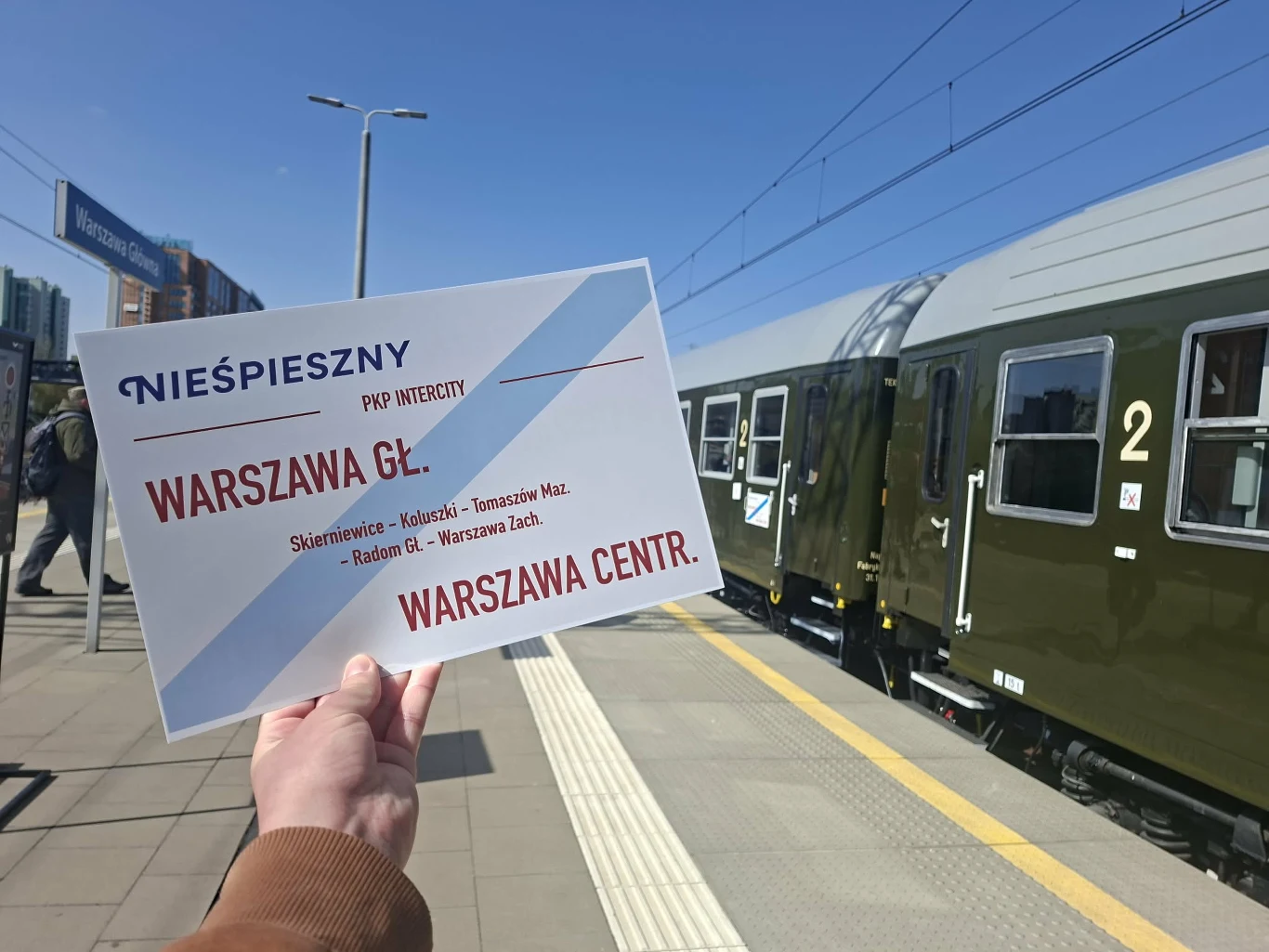 tablica kierunkowa z napisem 'Warszawa Gł.' i 'Warszawa Centr.' trzymana przez osobę na peronie, obok pociąg z oznaczeniem drugiej klasy, na tle słonecznego, bezchmurnego nieba