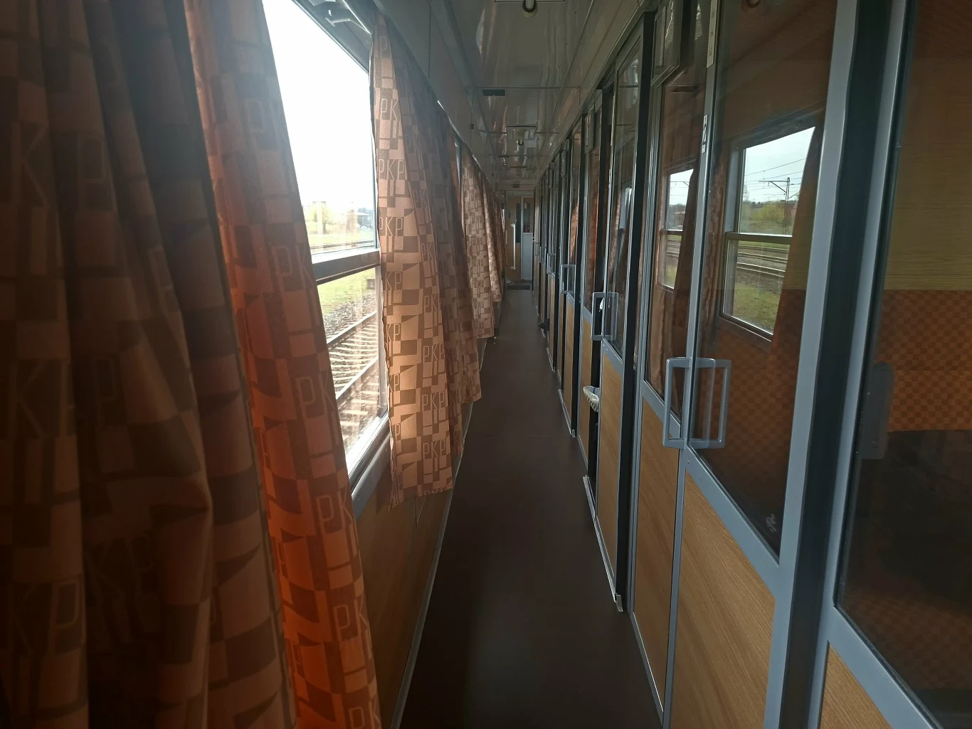 wąski korytarz wagonu kolejowego z dużymi oknami po lewej stronie, przez które wpada światło dzienne, oraz przeszklonymi drzwiami do przedziałów po prawej; na oknach widoczne brązowe zasłony z geometrycznym wzorem