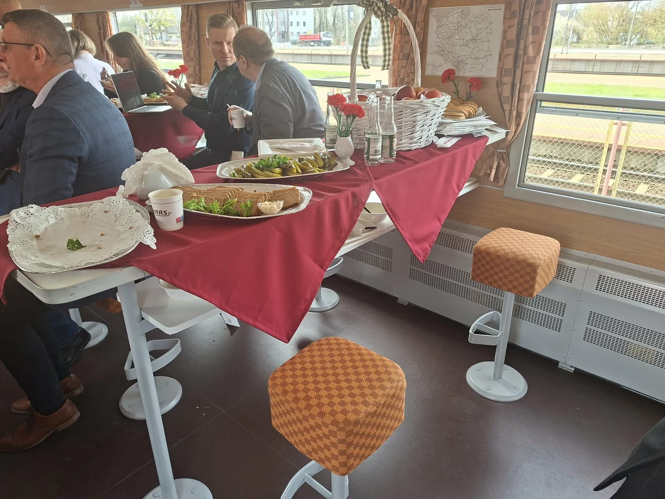 Grupa osób siedząca przy długim stole z czerwonym obrusem w wagonie restauracyjnym, na stole talerze z jedzeniem, półmiski z przystawkami i kwiaty w wazonach, w tle okno z widokiem na peron.