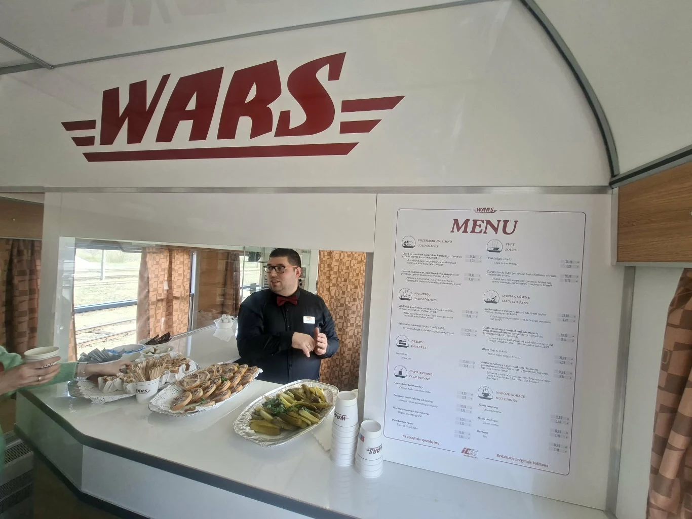 pracownik baru WARS w uniformie stoi za ladą, na której ustawione są tace z różnymi przekąskami i kubkami, obok widoczna wyraźnie karta menu