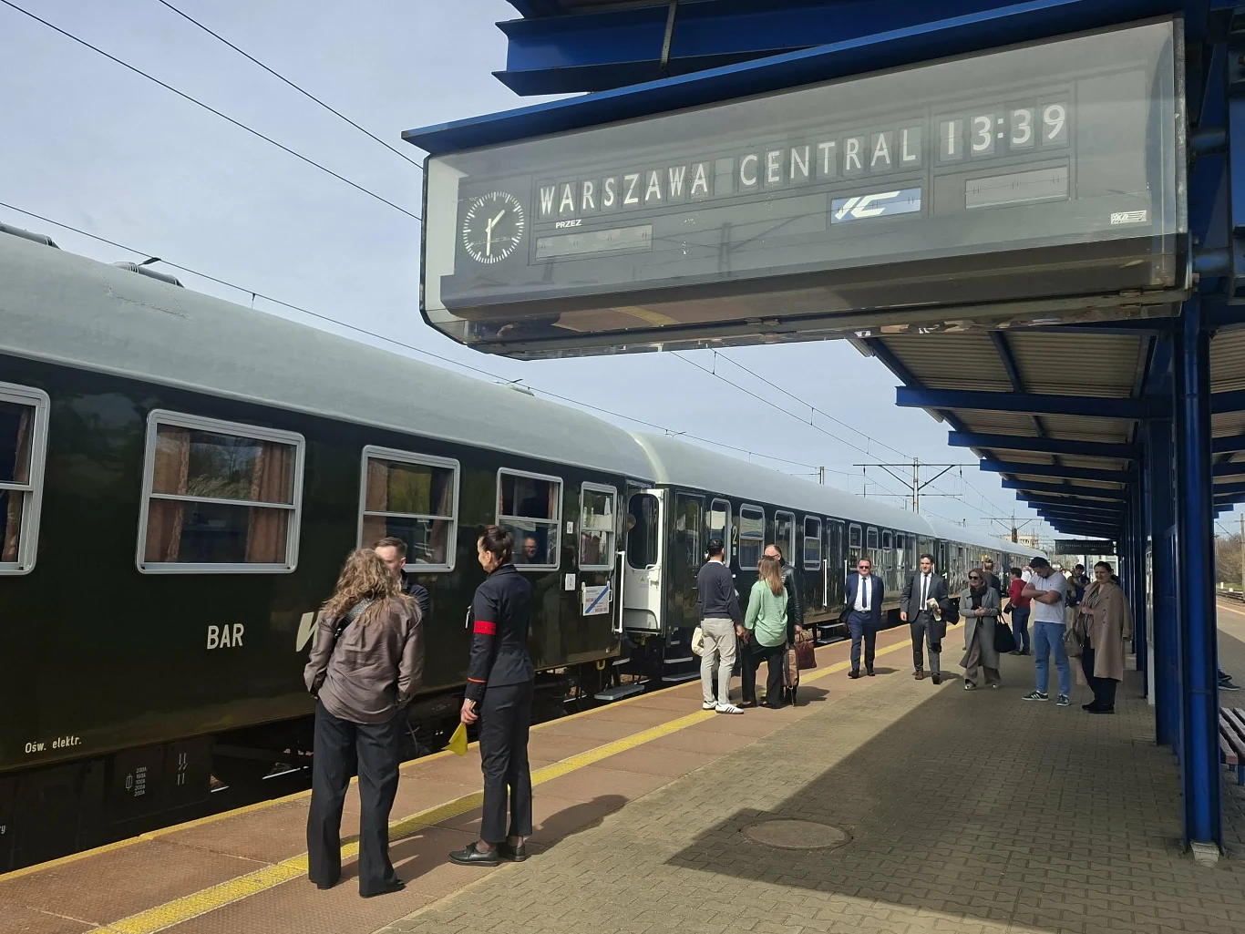 peron kolejowy z kilkunastoma osobami czekającymi przy zielonym wagonie pociągu, nad którym widoczna jest tablica z informacją o odjeździe do Warszawy Centralnej o godzinie 13:39