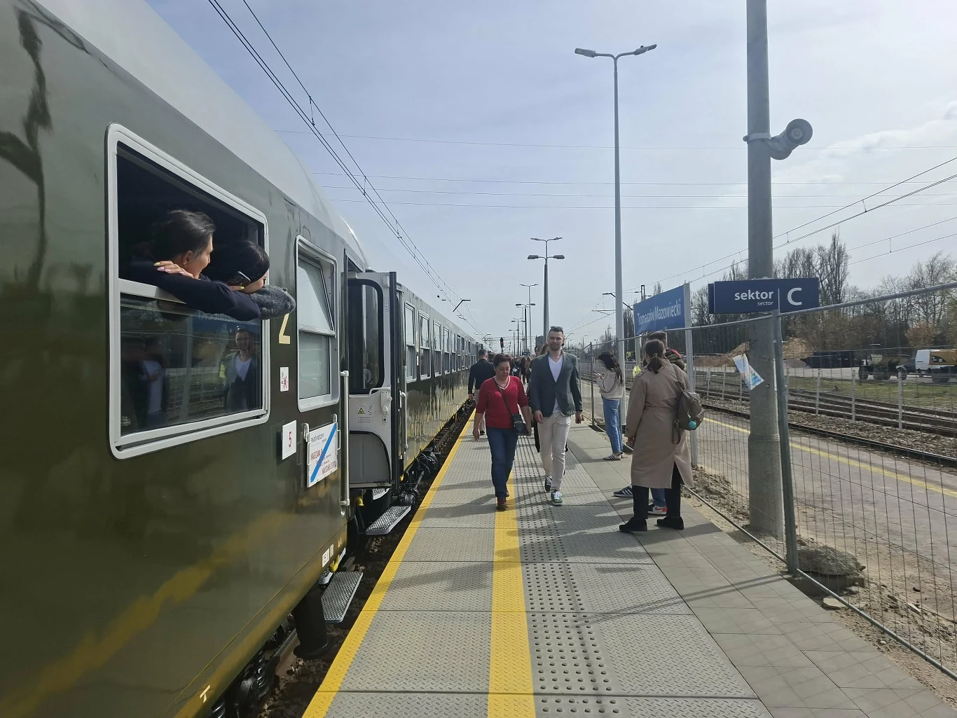 Peron z żółtą linią bezpieczeństwa, stojący pociąg z otwartymi oknami, przez które wyglądają pasażerowie, kilku ludzi z walizkami idących w stronę wyjścia oraz znak informujący o sektorze C.