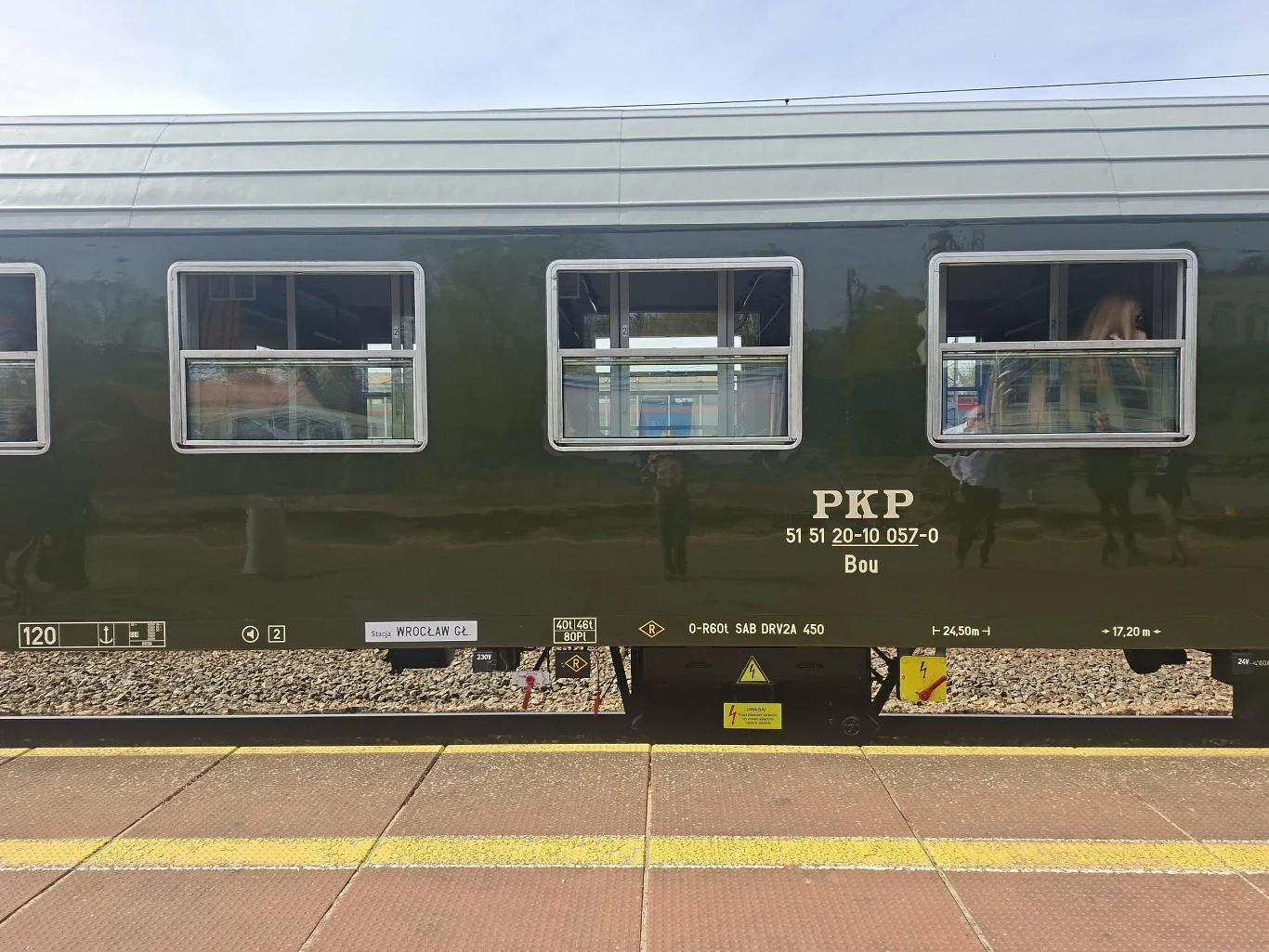 zielony wagon kolejowy PKP z otwartymi oknami, widocznymi napisami technicznymi i tabliczkami informacyjnymi, stojący przy peronie na tle torów