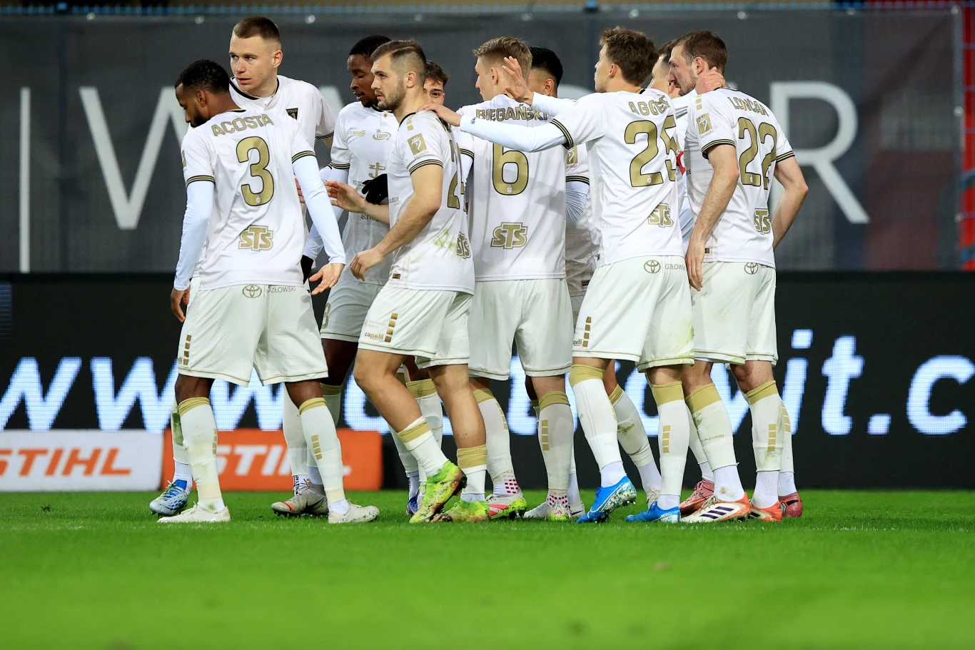 grupa piłkarzy w białych strojach z numerami i nazwiskami na plecach stoi razem na murawie stadionu, skupiona i zgrupowana po akcji meczowej, na pierwszym planie widoczne są numery zawodników oraz detale strojów sportowych