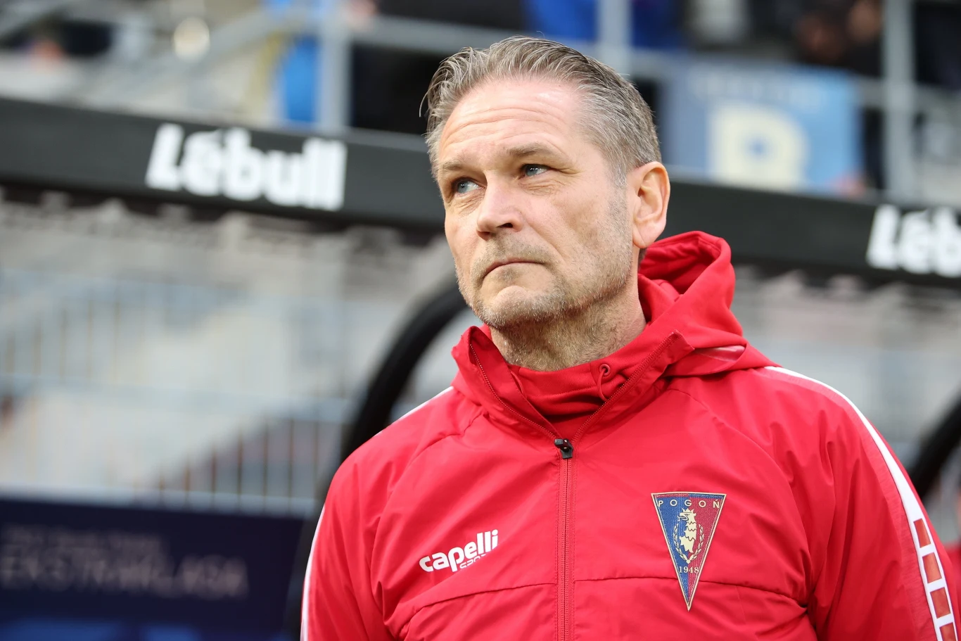Mężczyzna w średnim wieku w czerwonej kurtce sportowej z herbem klubu piłkarskiego Pogoń Szczecin stoi na tle stadionu, z poważnym wyrazem twarzy, skupiony i zamyślony.