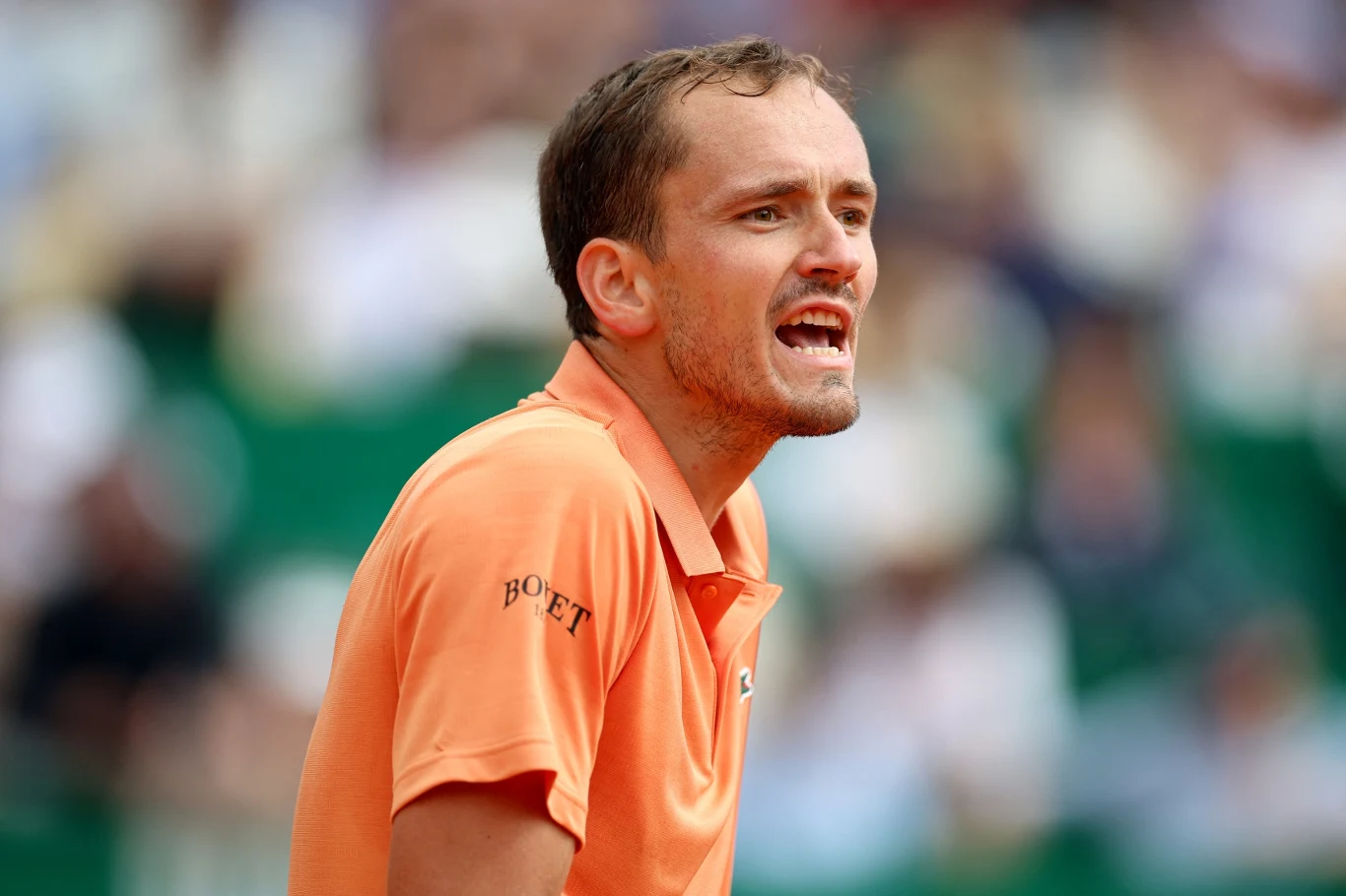 Daniił Miedwiediew Mężczyzna w pomarańczowej koszulce sportowej o krótkich rękawach wyraża silne emocje, patrząc w bok; w tle rozmyte sylwetki widzów na trybunach.