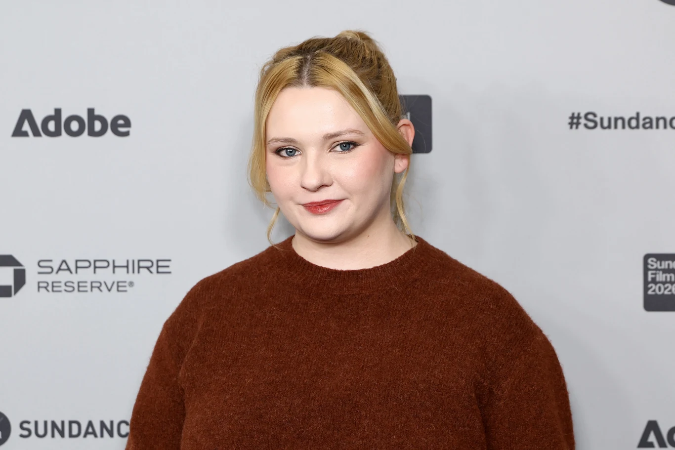 Abigail Breslin Kobieta z blond włosami spiętymi w kucyk, w bordowym swetrze, stoi na tle ścianki z logotypami firm oraz napisem 'Sundance Film Festival'.