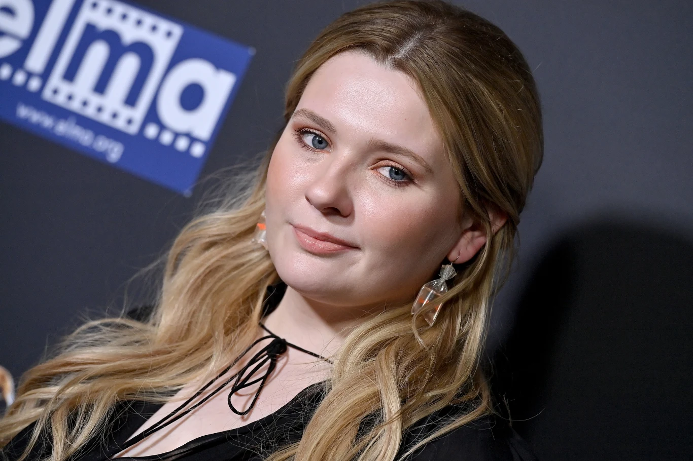 Abigail Breslin Abigail Breslin w czarnej sukience z wiązaniem pod szyją, blond włosy, jasna cera, geometryczne kolczyki, granatowe tło z logotypem