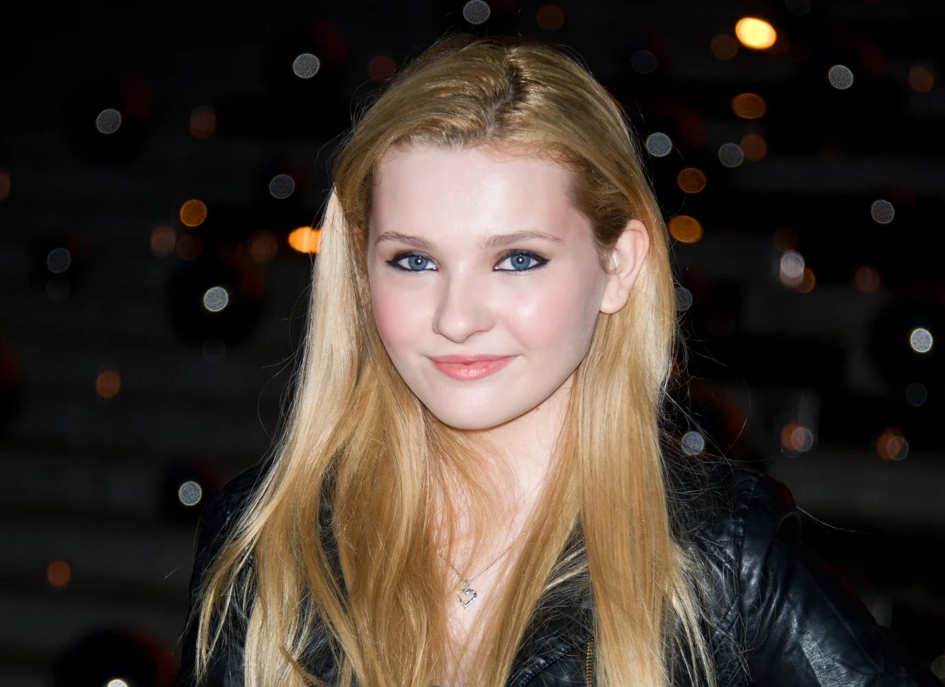 Abigail Breslin młoda kobieta z długimi blond włosami ubrana w czarną skórzaną kurtkę, delikatny makijaż podkreślający oczy, subtelny uśmiech, na szyi srebrny naszyjnik z zawieszką
