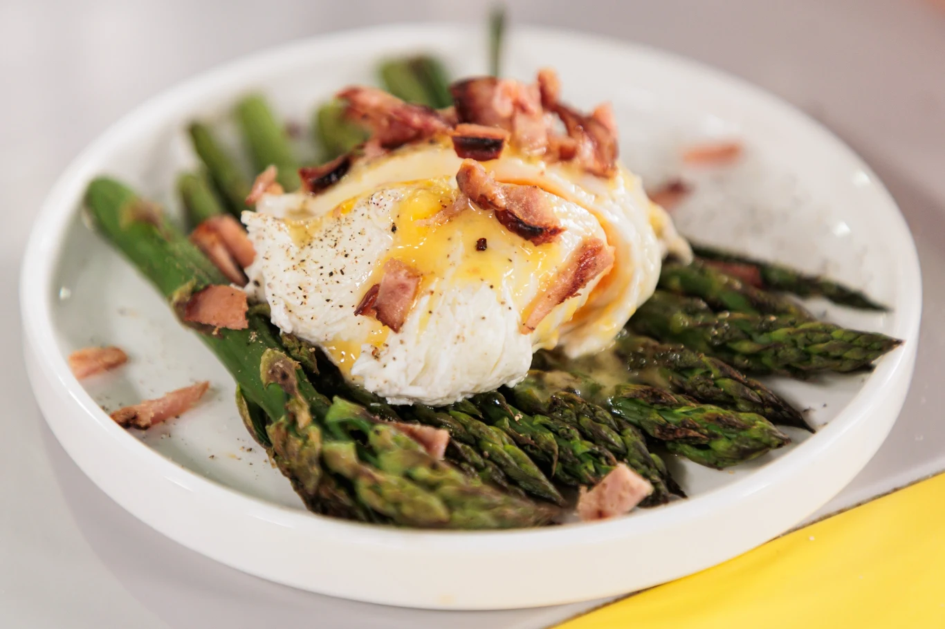 zielone szparagi ułożone w wachlarz, na nich jajko w koszulce z płynnym żółtkiem i kawałkami chrupiącego boczku, całość posypana świeżo zmielonym czarnym pieprzem