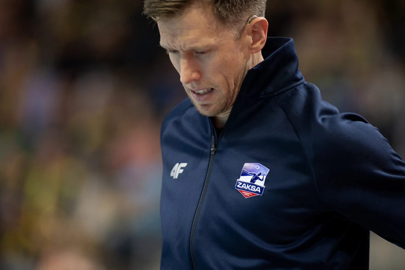 David Smith był związany z ZAKSĄ Kędzierzyn-Koźle Mężczyzna w granatowej kurtce sportowej z logo klubu siatkarskiego ZAKSA, pochylony w zamyśleniu, na rozmytym tle hali sportowej.