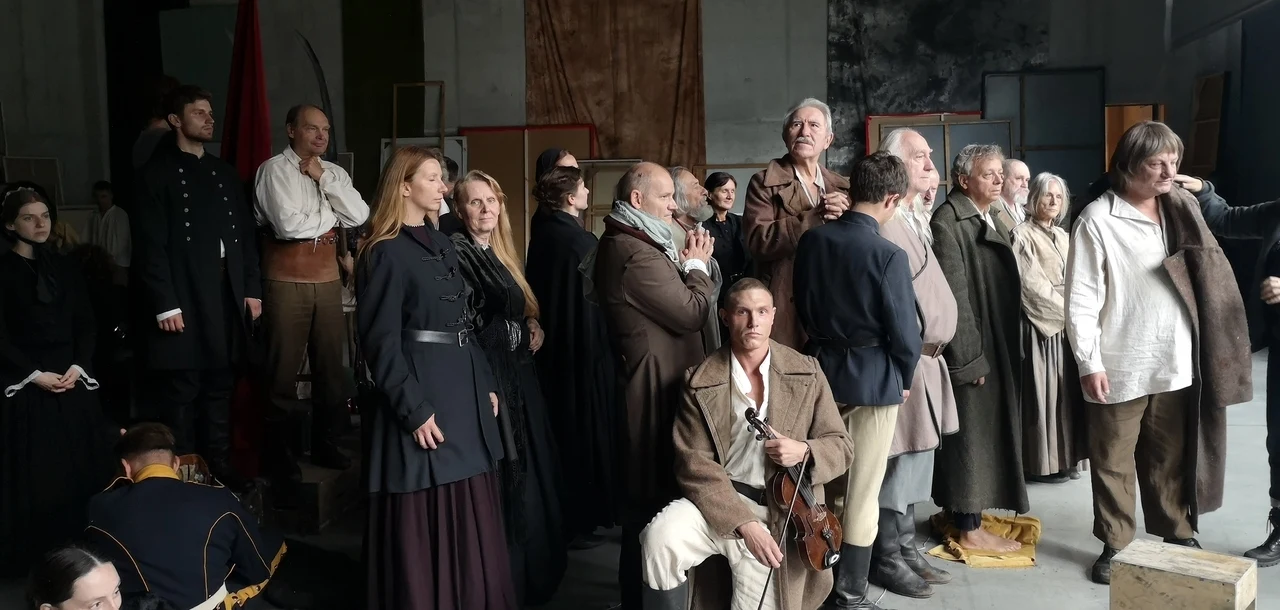 "12 obrazów zniewolenia" Lecha Majewskiego Grupa ludzi w historycznych strojach, część z nich ubrana w ciemne surduty i długie płaszcze, inni w jasne koszule i proste płócienne ubrania; centralna postać siedzi z bronią na kolanach, inni stoją w napięciu i skupieniu, jakby czekali na ważne wydar...