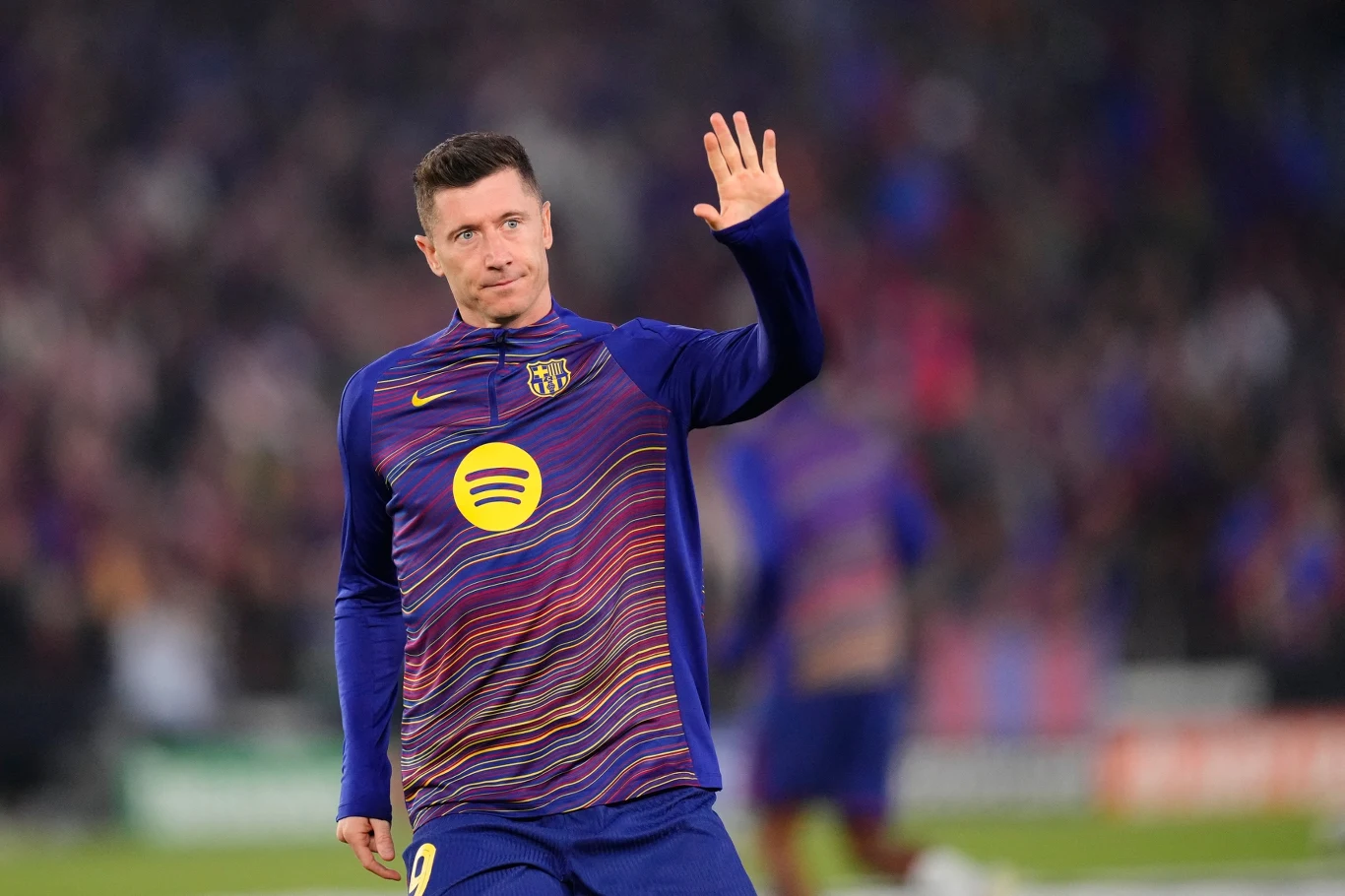 Piłkarz w granatowo-bordowej rozgrzewkowej bluzie FC Barcelony z logo Spotify na klatce piersiowej unosi dłoń w geście powitania, będąc na murawie stadionu; w tle rozmyte postacie i trybuny z kibicami.