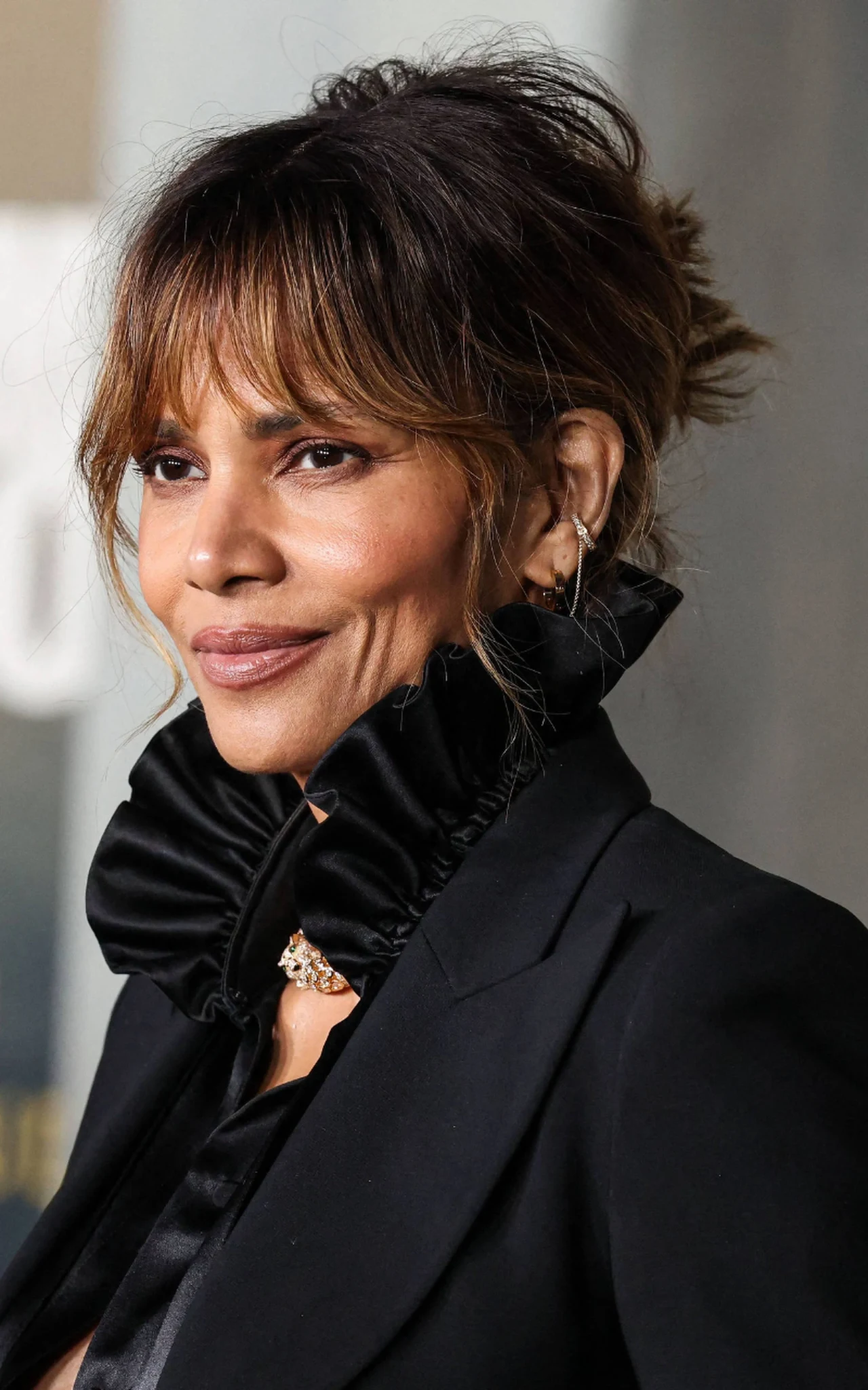 Halle Berry wyglądała świetnie w tej grzywce kobieta z upiętymi włosami i grzywką, ubrana w czarną marynarkę oraz bluzkę z wysokim, ozdobnym kołnierzem, delikatny makijaż podkreśla jej rysy twarzy