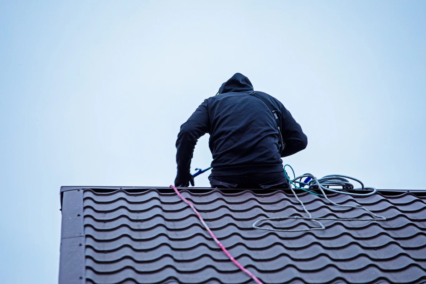Osoba w ciemnej kurtce pracująca na stromym, nowoczesnym dachu pokrytym metalowymi dachówkami, z liną zabezpieczającą i sprzętem do wspinaczki lub konserwacji.