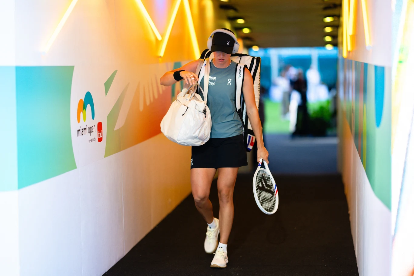 tenisistka w sportowym stroju idzie tunelem z torbą na ramieniu i rakietą tenisową w ręce, w tle rozmyte oświetlenie oraz logo turnieju Miami Open na ścianie