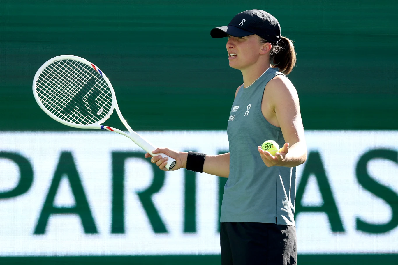 Tenisistka ubrana w sportowy top i czapkę z daszkiem stoi na korcie tenisowym, w jednej ręce trzyma rakietę, a w drugiej piłkę tenisową, wykonując nieco zdezorientowany gest dłonią.