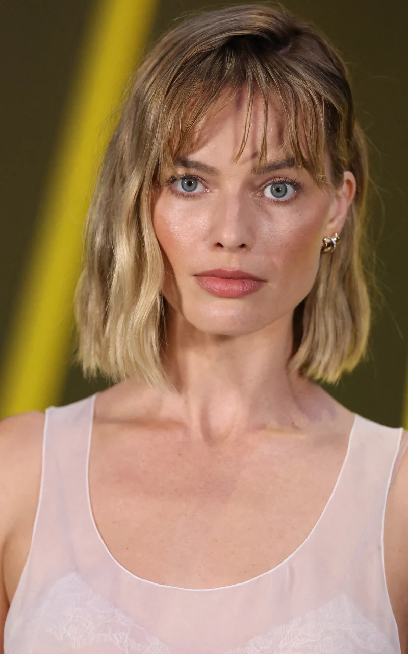 Margot Robbie zrobiła sobie cienką, lekką grzywkę i wygląda świetnie Kobieta o blond, krótkich, falowanych włosach i niebieskich oczach w delikatnym makijażu, ubrana w jasną sukienkę na ramiączkach, spogląda prosto w obiektyw na tle z żółtym, rozmytym akcentem.
