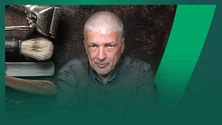 Starszy mężczyzna z siwymi włosami i krótkim zarostem patrzy pewnie wprost na obiektyw, w tle widoczne są narzędzia fryzjerskie, takie jak maszynka, pędzel i brzytwa, które podkreślają motyw barberski zdjęcia.