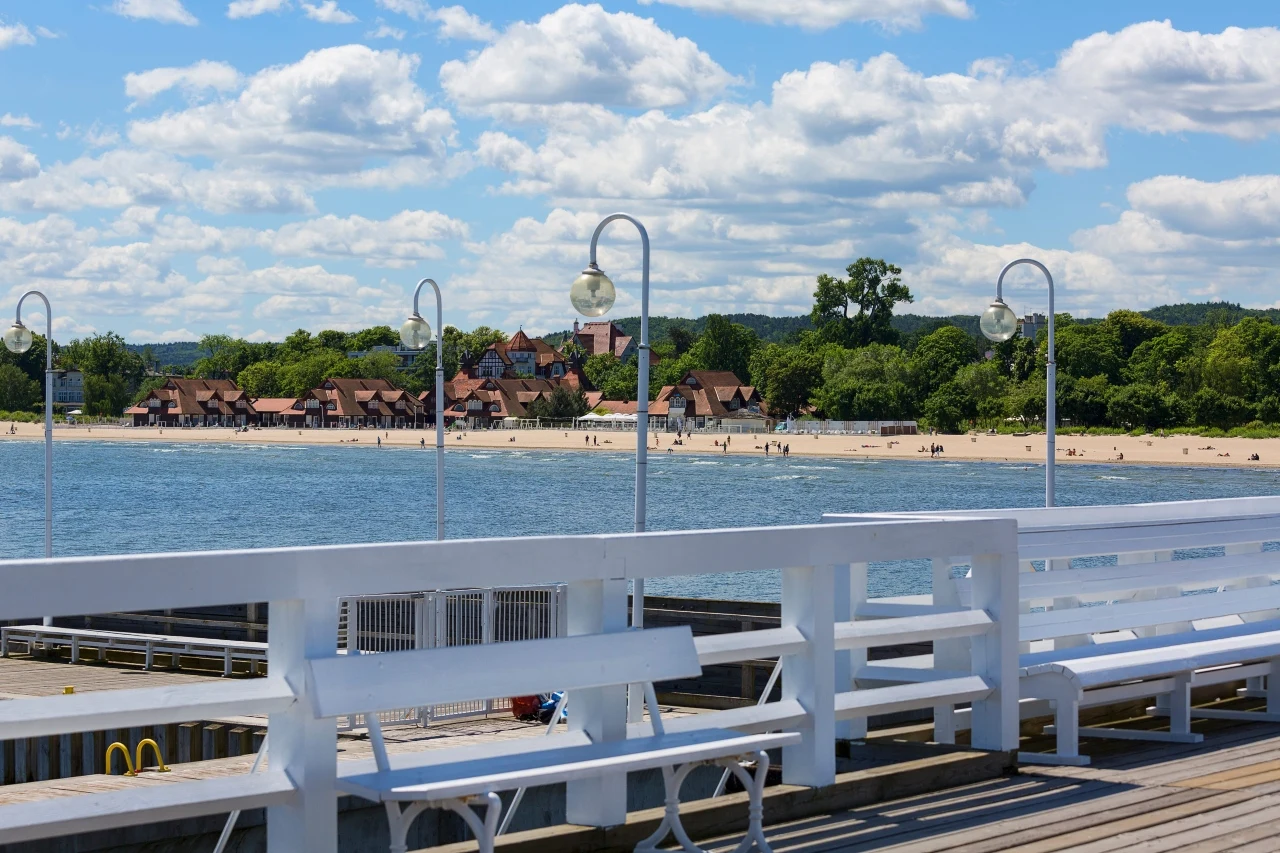 drewniane molo z białymi ławkami i latarniami, widok na piaszczystą plażę i rząd niskich budynków otoczonych zielenią po drugiej stronie wody, pod błękitnym niebem z licznymi chmurami