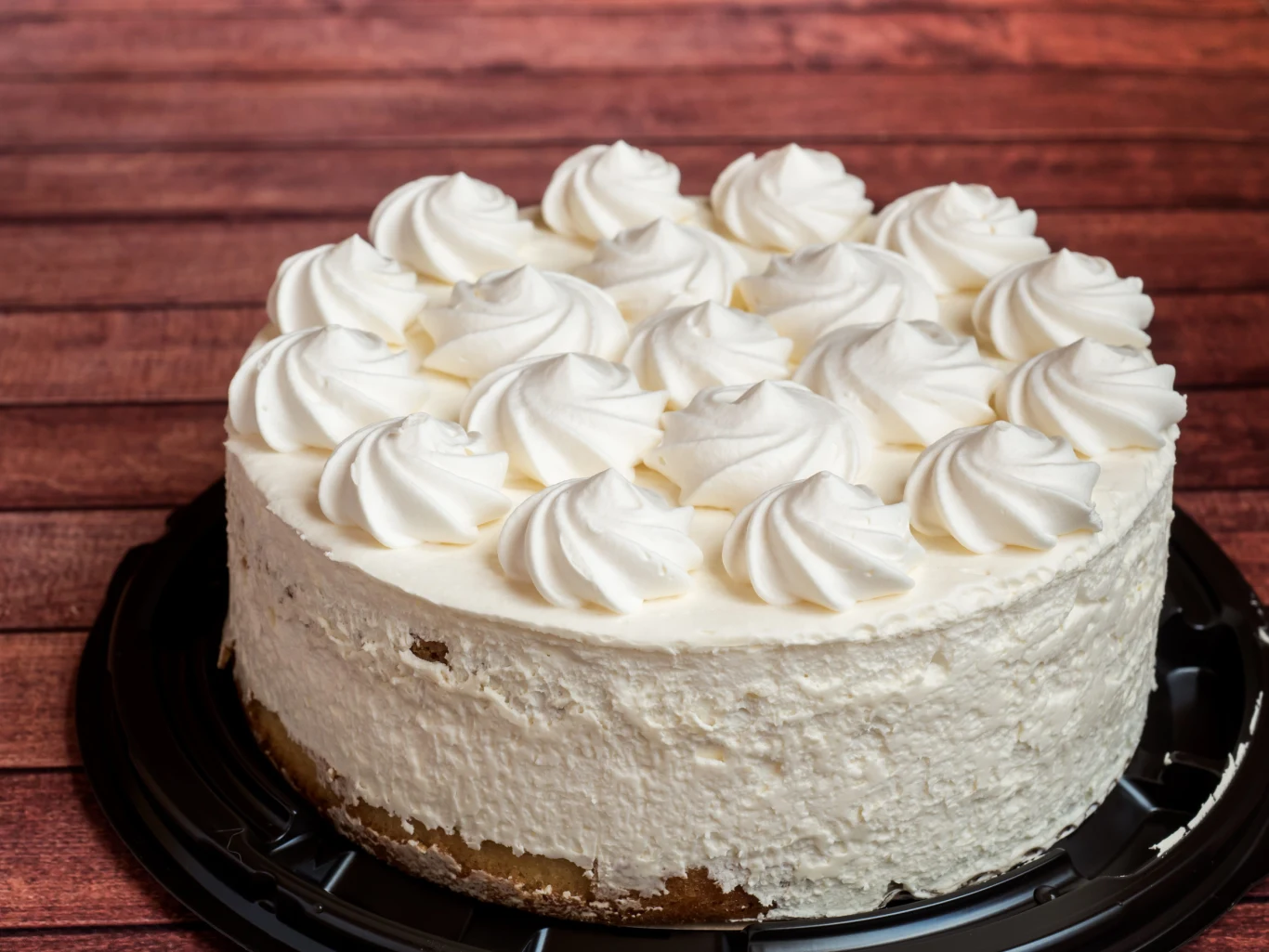 Poznaj sprawdzony przepis na prosty tort na Pierwszą Komunię Świętą Okrągły tort komunijny z bitą śmietaną i rozetkami z kremu na czarnym talerzu, na tle drewnianych paneli