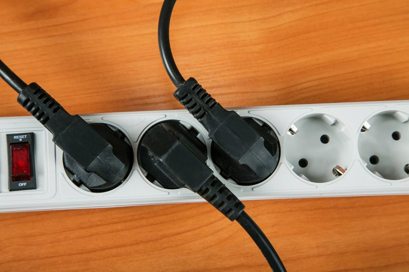 Biała listwa zasilająca z trzema podłączonymi czarnymi wtyczkami, widocznym czerwonym wyłącznikiem oraz dwoma wolnymi gniazdami, umieszczona na drewnianym blacie.