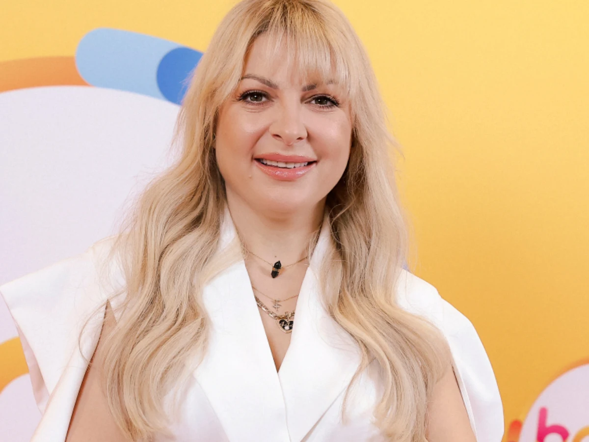 Magda Narożna Magda Narożna w białej stylizacji, długie blond włosy i grzywka, dwa naszyjniki, kolorowe tło żółto-niebiesko-pomarańczowe.