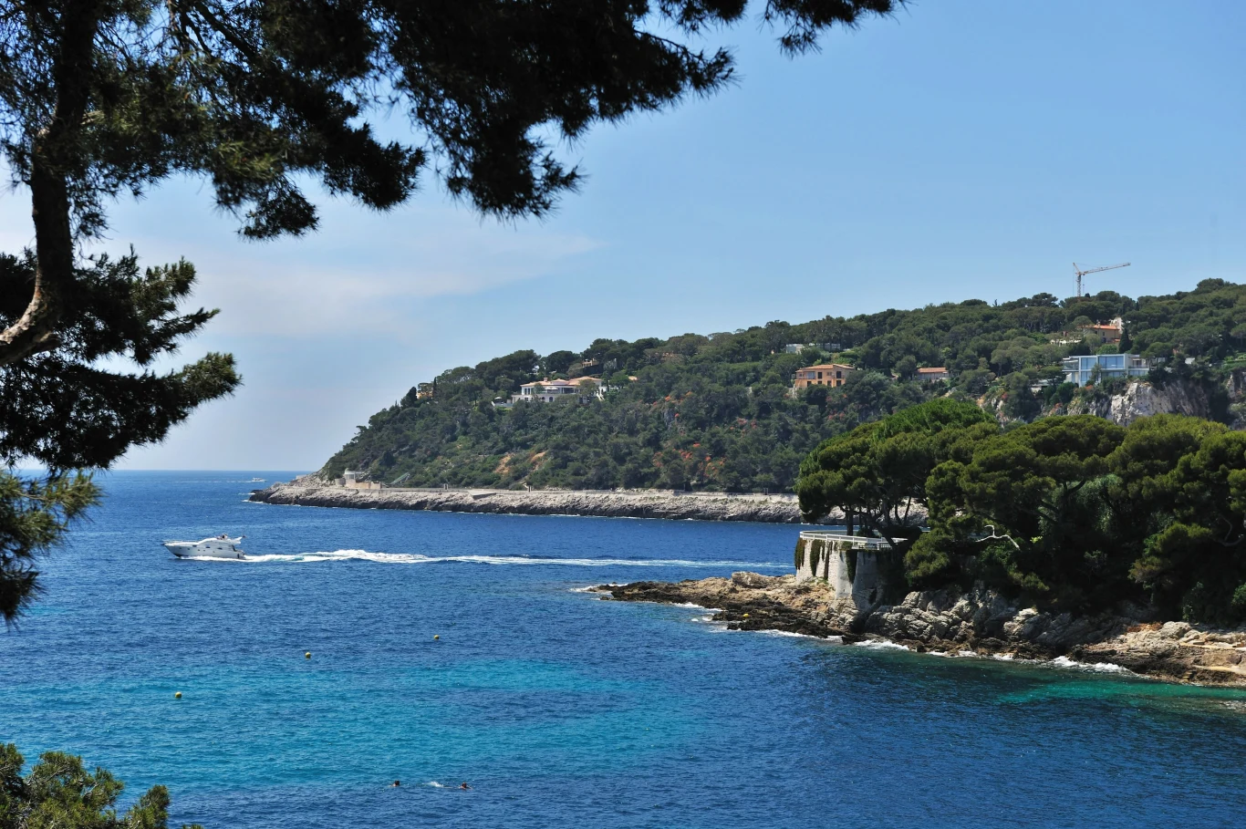 Monako zachwyca swoim malowniczym położeniem. Błękitne morze oblewające skaliste wybrzeże porośnięte zielonymi drzewami i willami, szybka łódź motorowa płynąca blisko brzegu, w tle łagodna linia wzgórza z pojedynczymi budynkami.