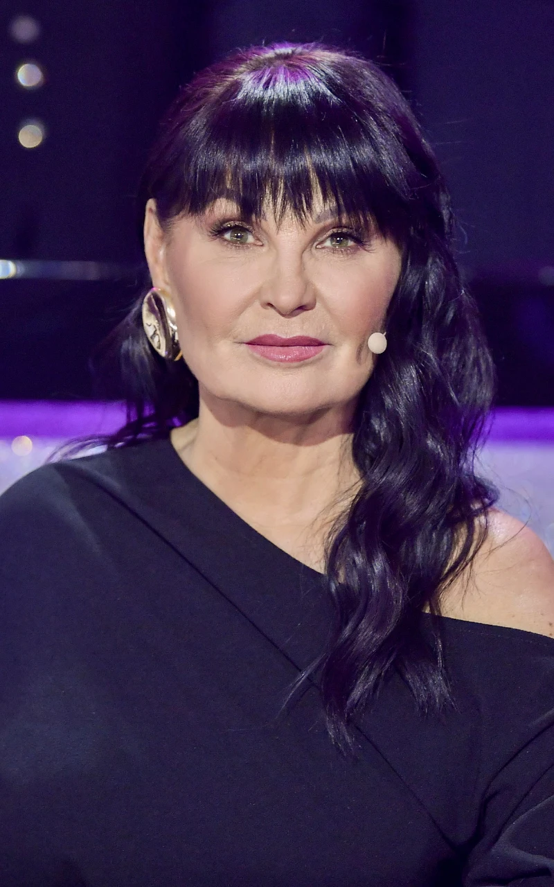 Iwona Pavlović i jej ikoniczna grzywka kobieta z długimi, czarnymi włosami ułożonymi w fale, ubrana w czarną bluzkę z odsłoniętym jednym ramieniem, z dużymi kolczykami i mikrofonem przy twarzy, na tle fioletowego światła studyjnego