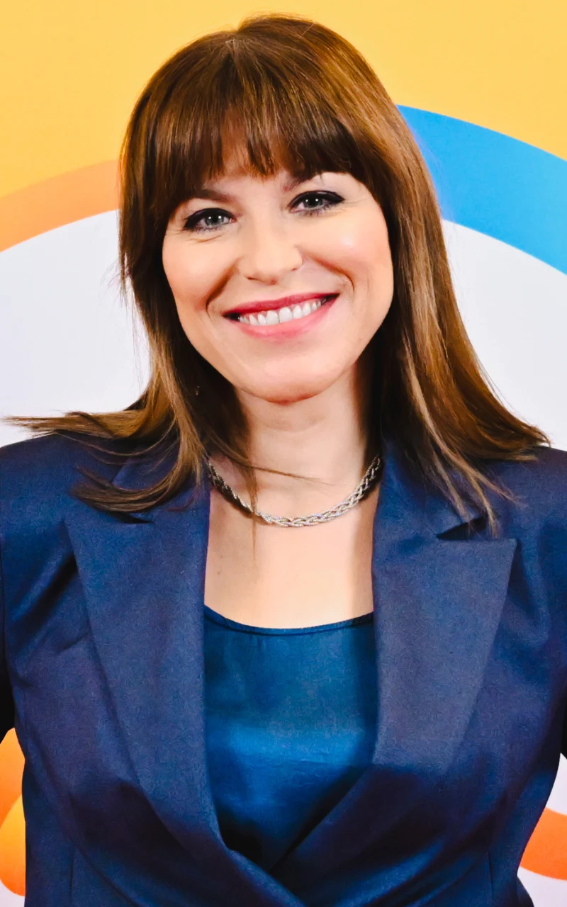 Katarzyna Bonda od niedawna jest dumną właścicielką grzywki uśmiechnięta kobieta z prostymi, brązowymi włosami i grzywką, ubrana w granatowy żakiet i top, na szyi srebrny łańcuszek, za nią kolorowe tło z żółtym i niebieskim okręgiem