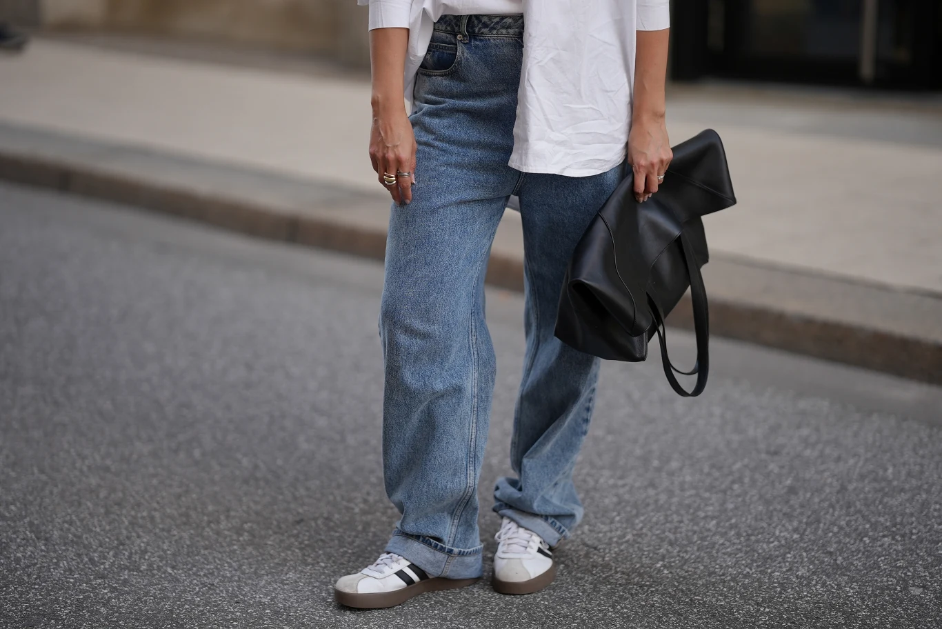 Flare jeans i sneakersy - jedno z najmodniejszych połączeń wiosny 2026 osoba ubrana w luźne niebieskie dżinsy i białe sneakersy trzyma dużą czarną kopertówkę, ubrana także w białą koszulkę o długich rękawach