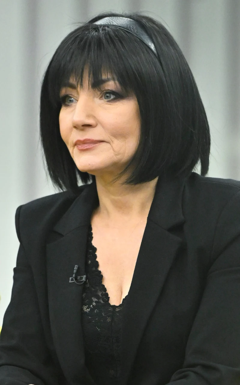 Shazza, ikona disco-polo, grzywkę nosi już od wielu lat Kobieta o ciemnych, prostych włosach z grzywką, ubrana w czarną marynarkę i koronkowy top, siedzi zwrócona lekko bokiem na jasnym, neutralnym tle.