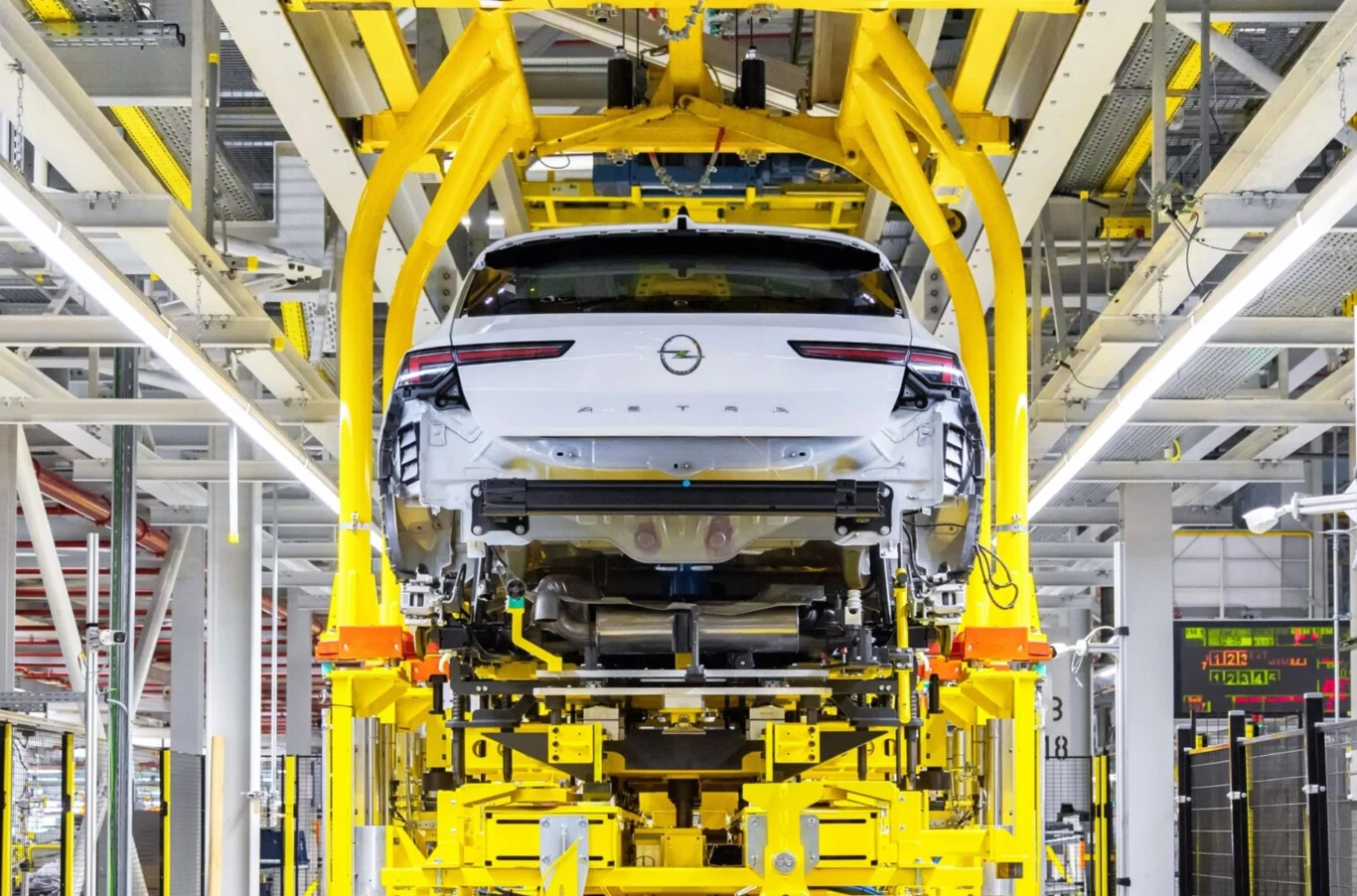 Kolejne cięcia w Oplu. Inżynierowie tracą pracę, zmienia się strategia. Biały Opel na linii montażowej w fabryce, otoczony żółtymi maszynami przemysłowymi.