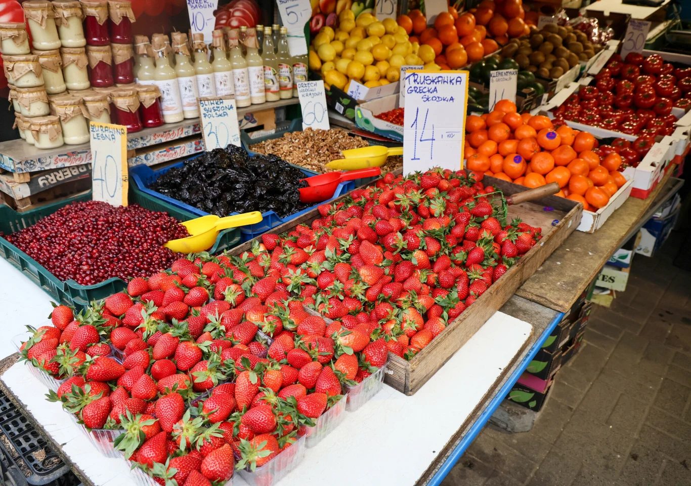 Greckie truskawki mają duże szypułki Greckie truskawki z dużymi szypułkami na straganie obok czereśni, śliwek, mandarynek, cytryn i słoików z przetworami.