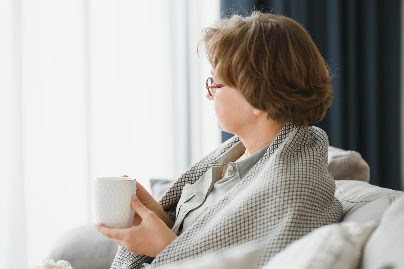 Dlaczego seniorom jest zimno nawet w ciepłym pomieszczeniu? Kobieta owinięta kocem siedzi na kanapie, trzymając w rękach biały kubek i patrzy w stronę jasnego okna.