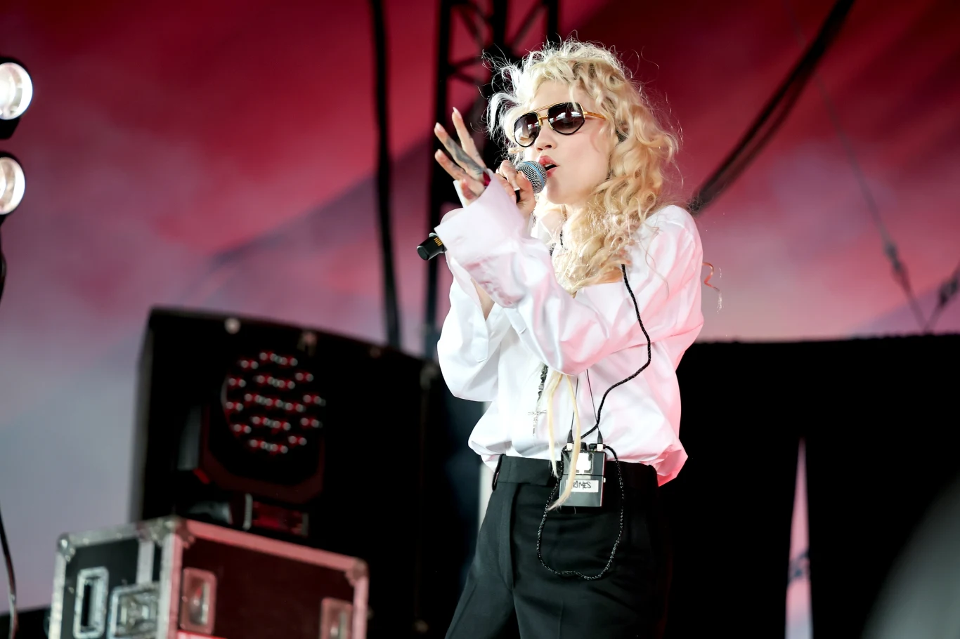 Grimes wystąpiła na Coachelli Wokalistka w okularach przeciwsłonecznych i białej koszuli śpiewa do mikrofonu na scenie koncertowej, otoczona sprzętem muzycznym i charakterystycznym oświetleniem.