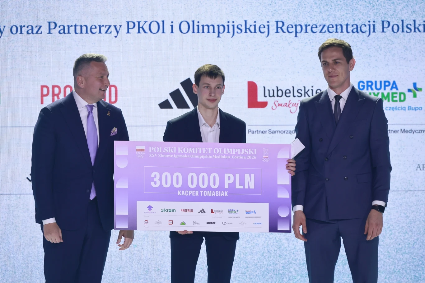 Kacper Tomasiak odebrał nagrody w siedzibie PKOl trzech mężczyzn ubranych w garnitury, środkowy trzyma wielki fioletowy czek na 300 000 PLN z widocznym napisem „Kacper Tomasiak”; w tle jasna ścianka z logotypami sponsorów i nazwiskami partnerów