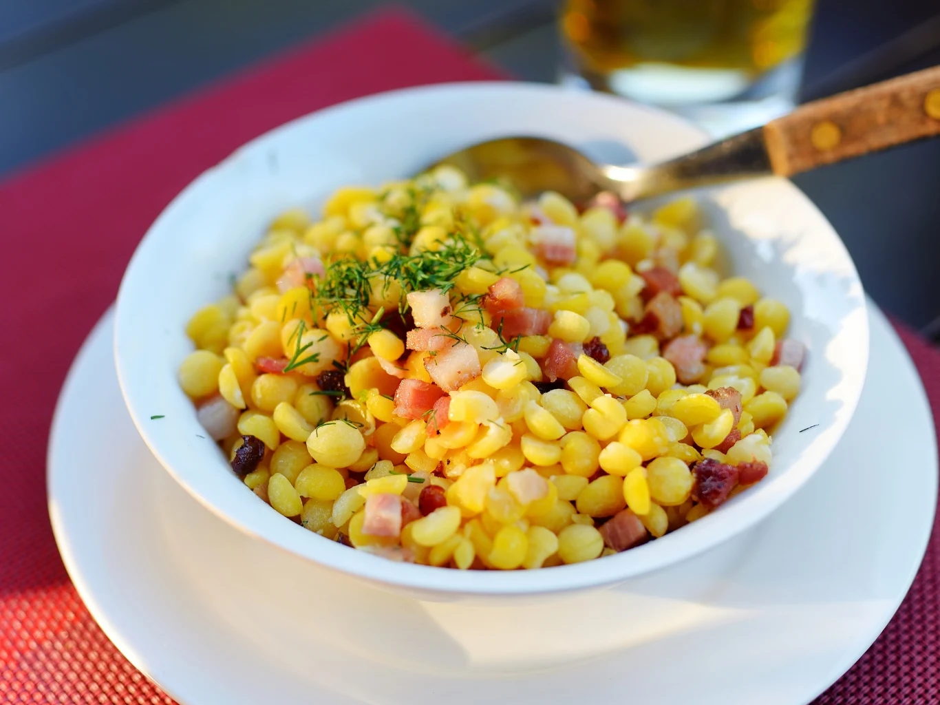 Groch łuskany to skarbnica białka i innych witamin. Można z niego zrobić nie tylko sycącą zupę. Biała miska z grochem łuskanym, boczkiem i koperkiem, obok łyżka z drewnianą rączką.