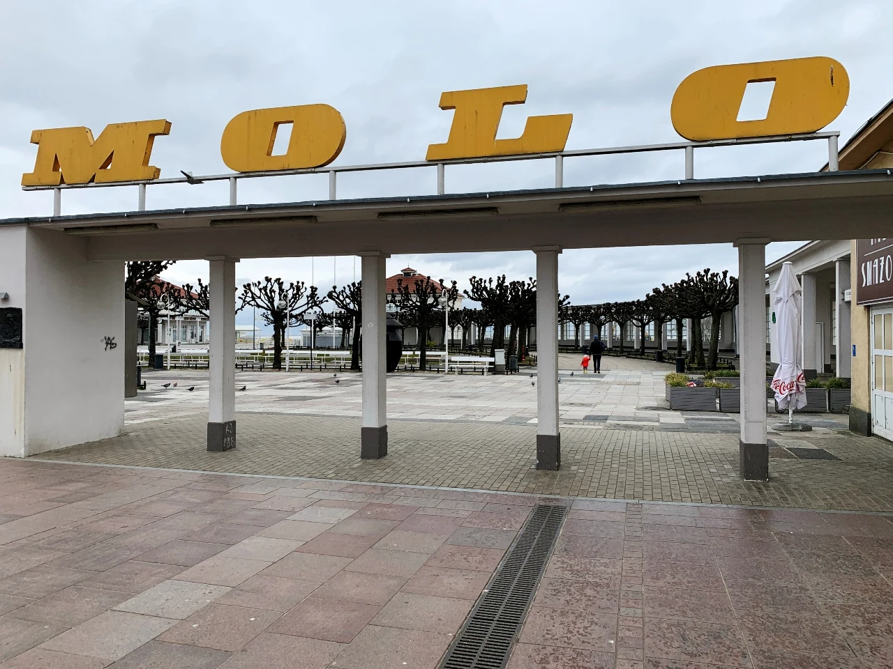 wejście na molo z dużym, żółtym napisem „MOLO” zamocowanym na białej konstrukcji, w tle rząd przyciętych drzew i widoczne zabudowania oraz pojedynczy przechodzień na deptaku