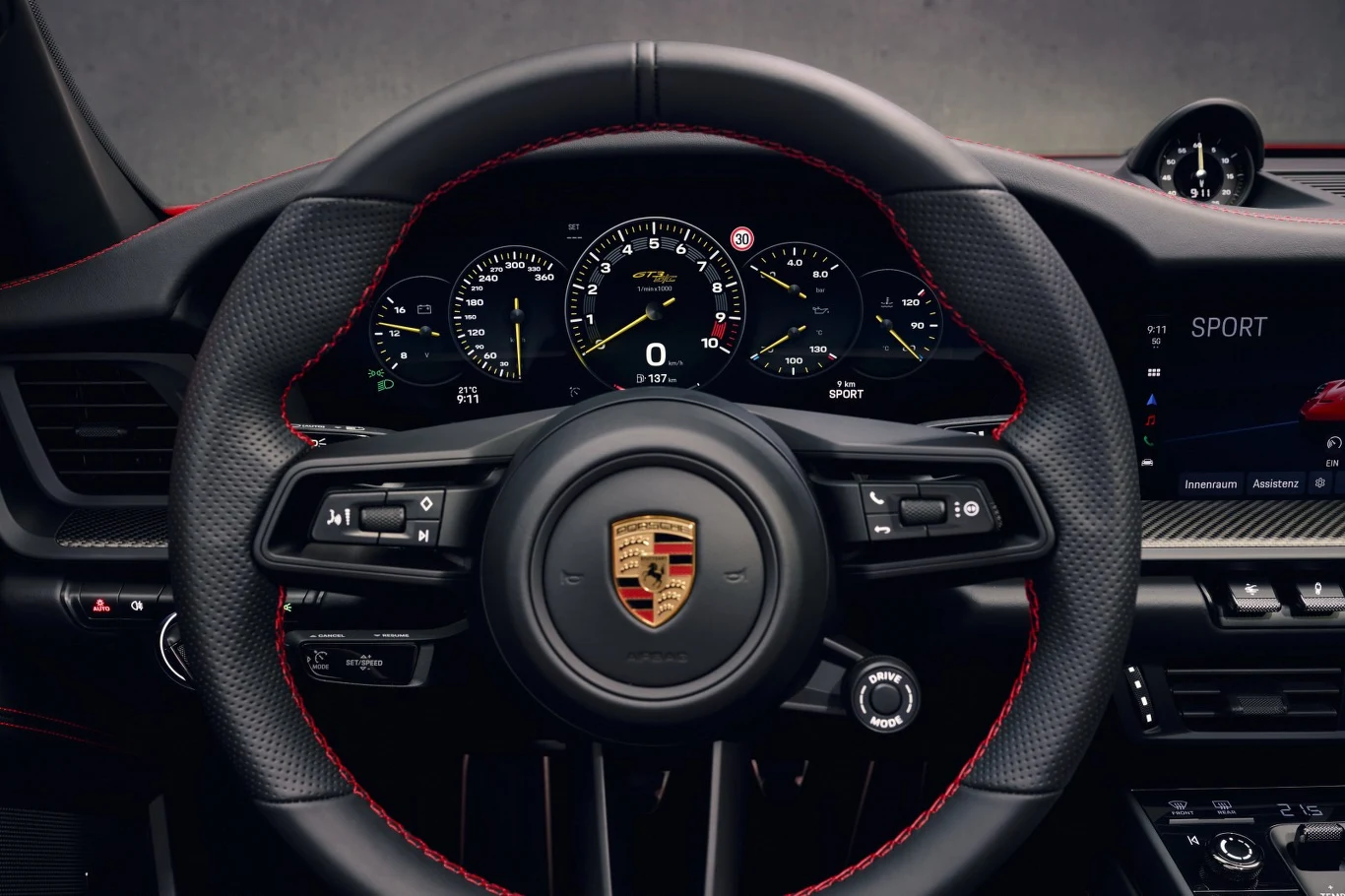 kierownica samochodu z logo Porsche, nowoczesny panel zegarów z podświetlanymi wskaźnikami oraz wyświetlaczami cyfrowymi, wnętrze wykończone sportowym charakterem, czerwone przeszycia na skórze