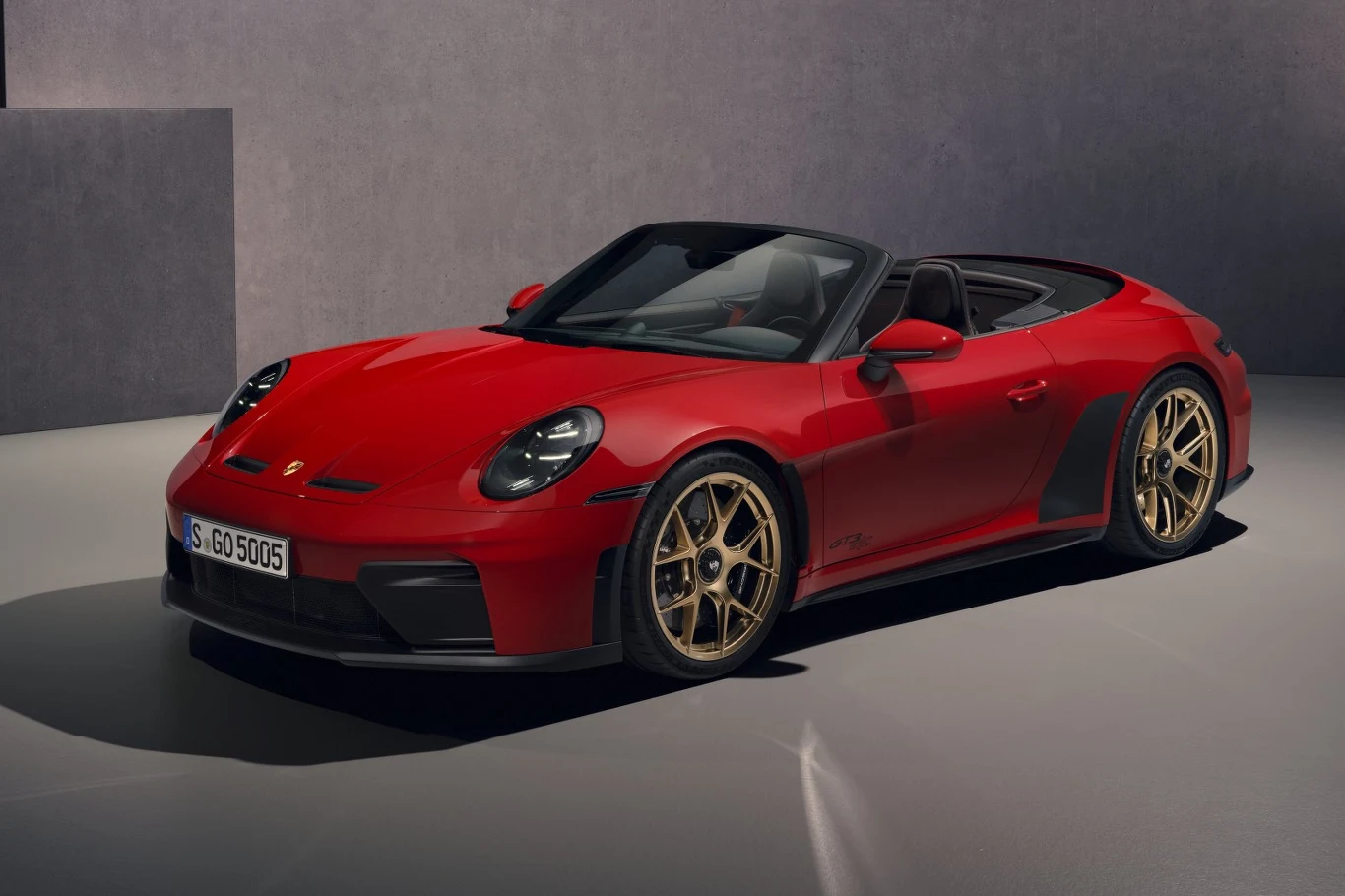 Czerwony kabriolet sportowy Porsche z otwartym dachem, złotymi felgami i aerodynamicznymi wstawkami na bokach, ustawiony na minimalistycznym tle we wnętrzu o neutralnych kolorach.