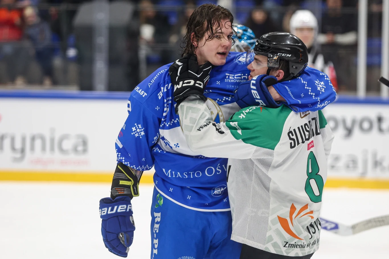 Roman Diukow Roman Diukow podczas meczu hokejowego; dwóch zawodników na lodowisku, kibice i bandy w tle.