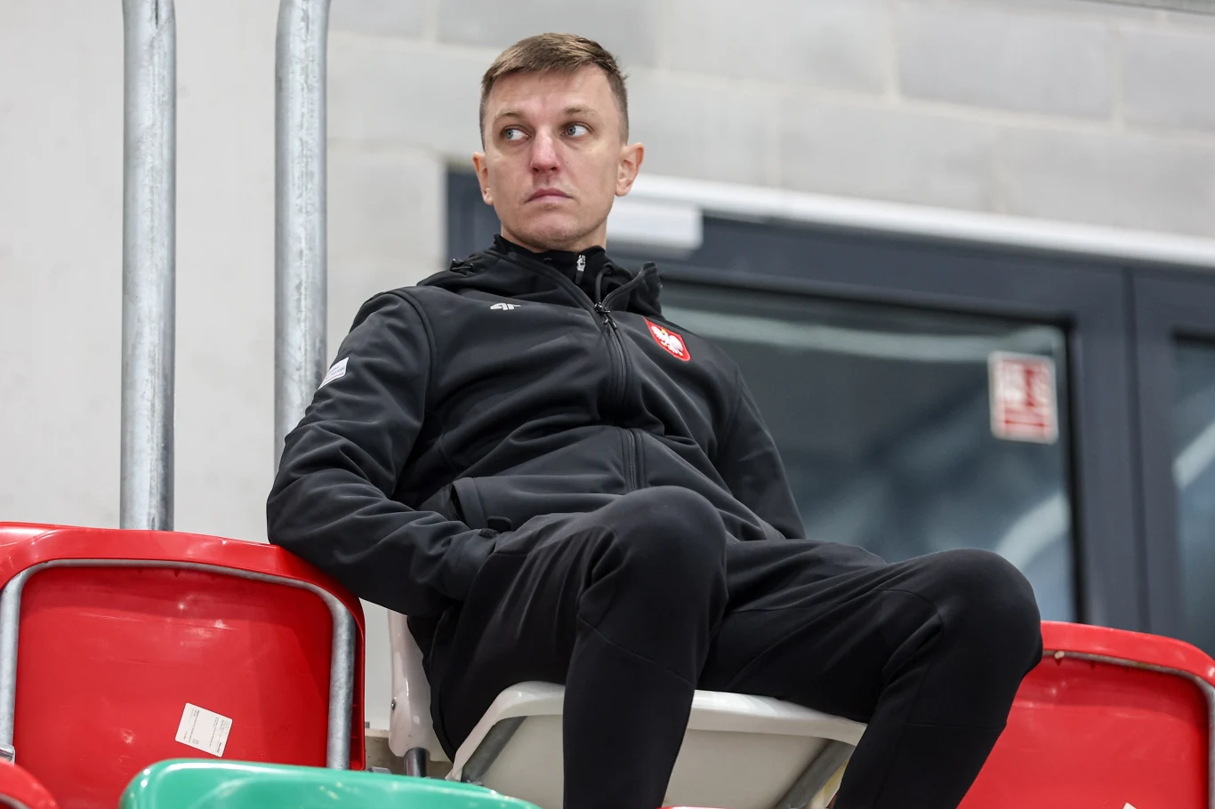 Jarosław Rzeszutko mężczyzna w czarnej sportowej kurtce z emblematem Polski siedzi samotnie na trybunach z czerwonymi i zielonymi krzesłami, patrząc w bok