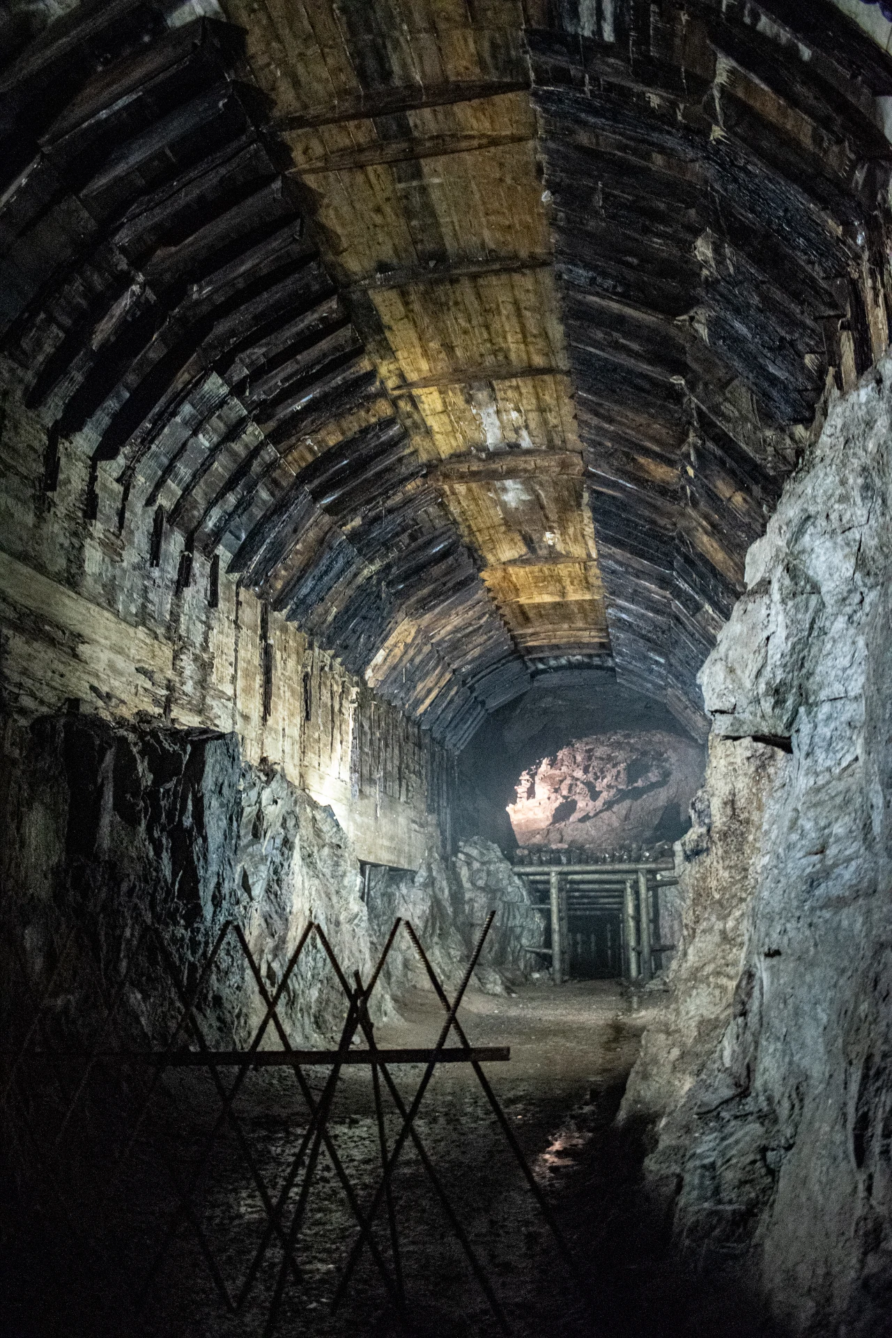"Podziemne miasto" Osówka. kamienno-betonowy tunel wykuty w skale, sklepienie wzmocnione drewnianymi belkami, w głębi widoczne zabezpieczenia i zamknięte barierki blokujące wejście dalej