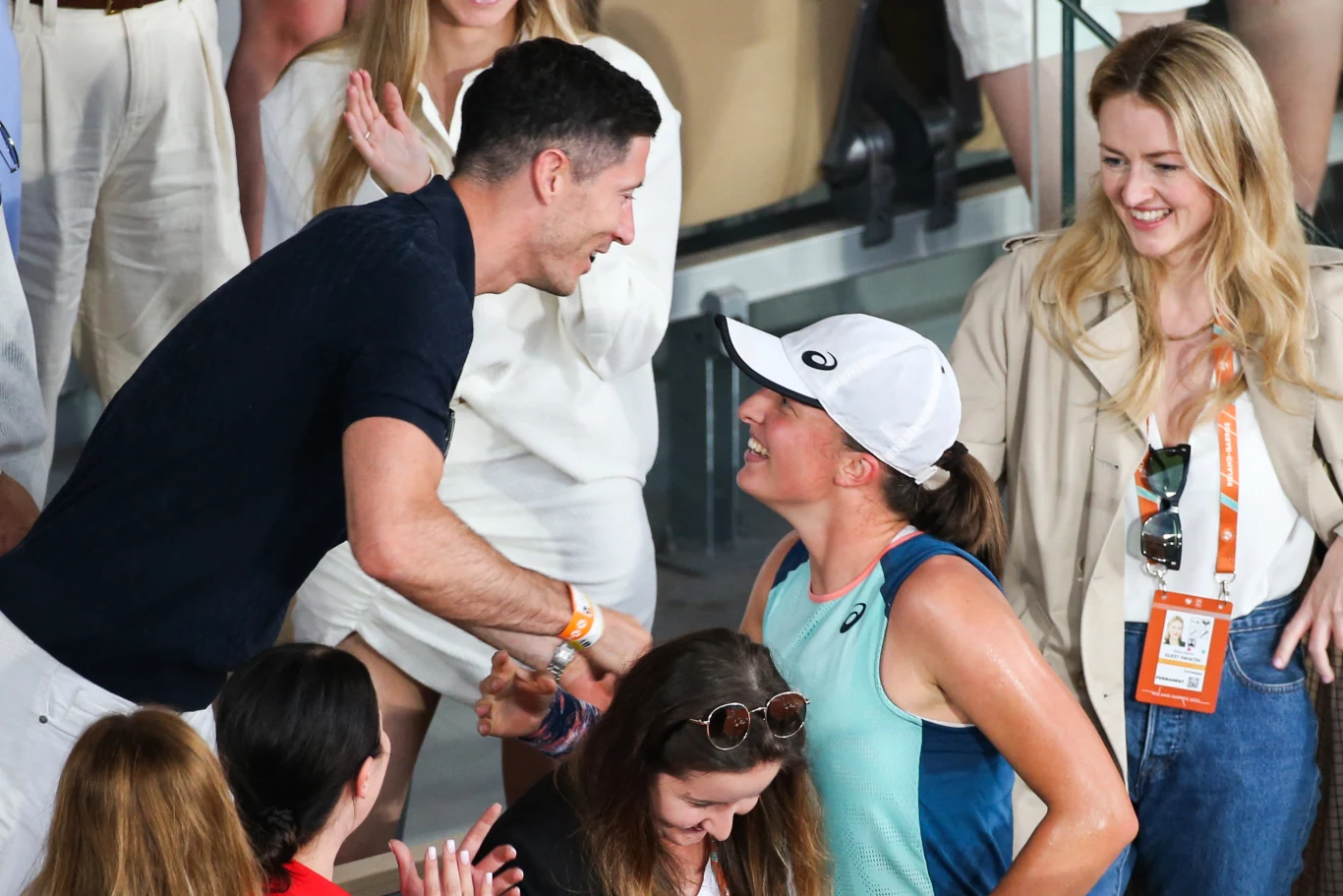 Mężczyzna w ciemnej koszulce serdecznie wita się z uśmiechniętą kobietą w sportowym stroju i białej czapce, obok młoda kobieta z identyfikatorem na szyi patrzy z radością na tę scenę; w tle grupa osób obserwujących spotkanie.